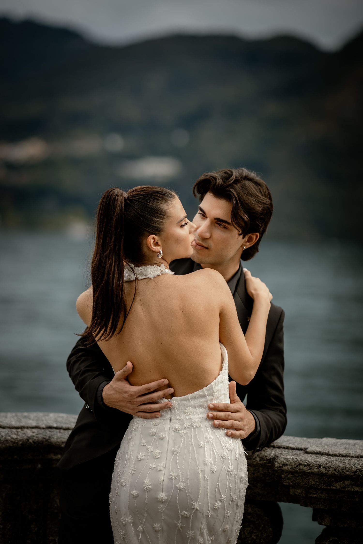 Romantic wedding couple embracing on a terrace overlooking a scenic mountain lake with cloudy skies.