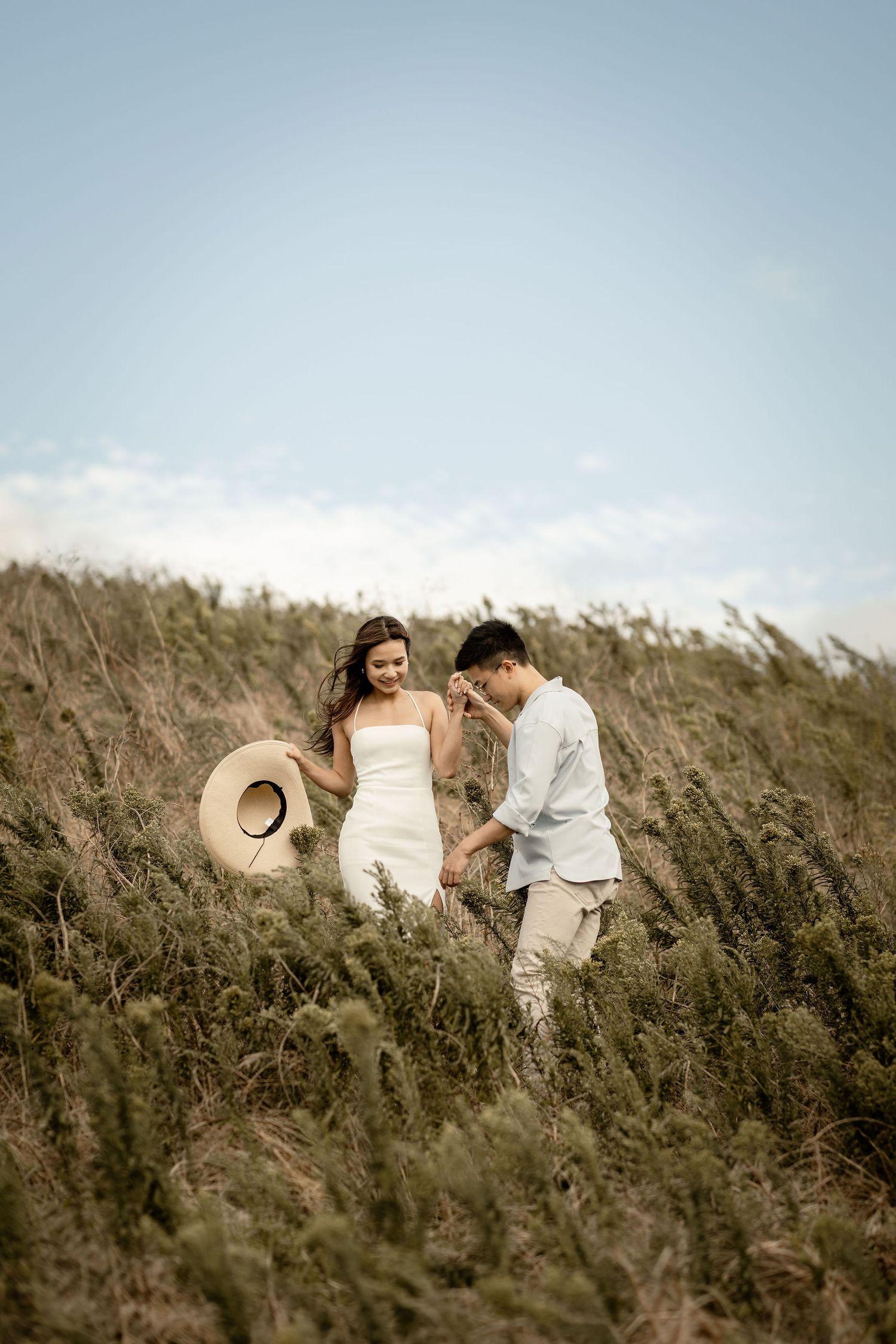 A couple stands together in tall grass against a blue sky.