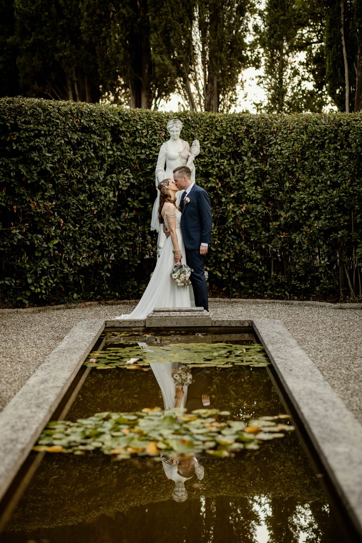 Wedding couple standing by reflecting pool with hedge backdrop in formal garden setting.