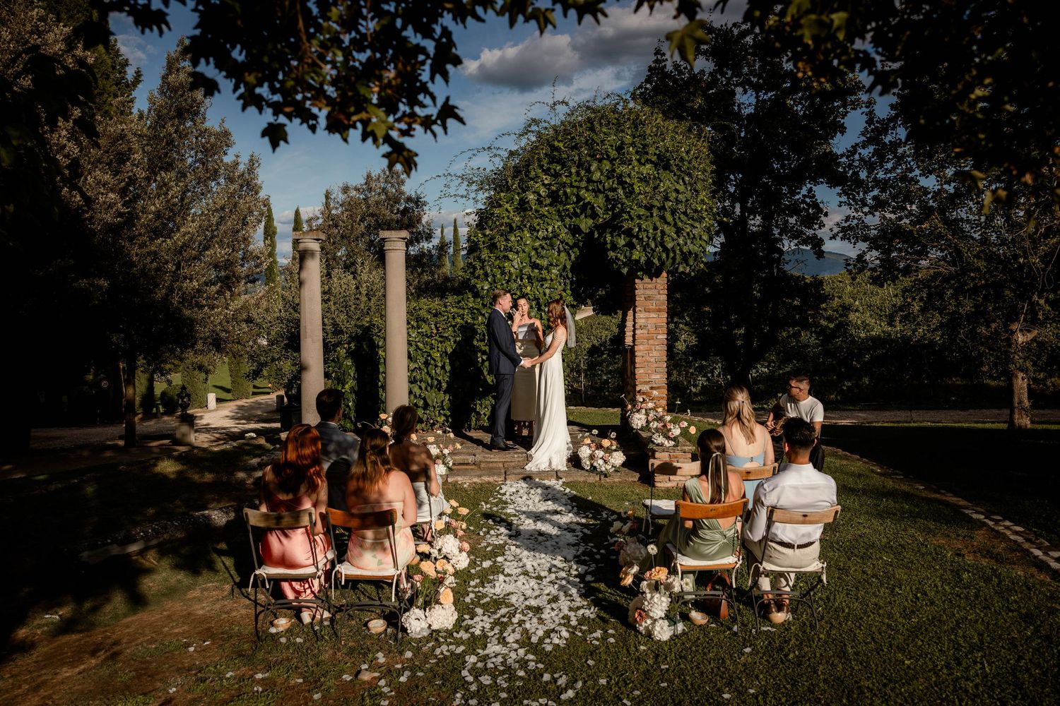 An intimate outdoor wedding ceremony takes place under stone pillars with guests seated on a flower-lined path.