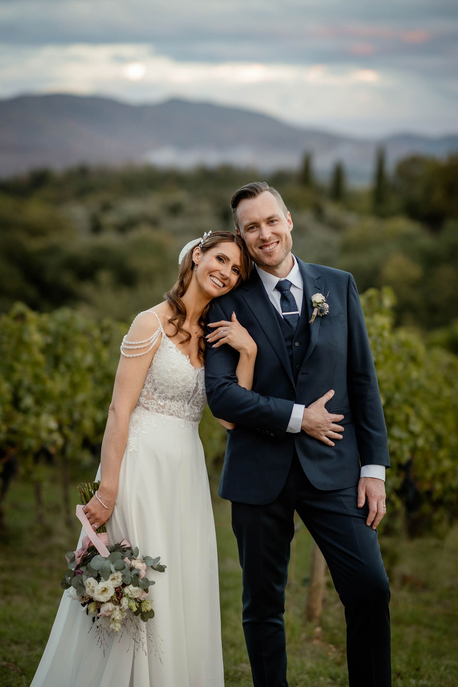Couple poses elegantly in vineyard setting with dramatic mountain vista behind them.