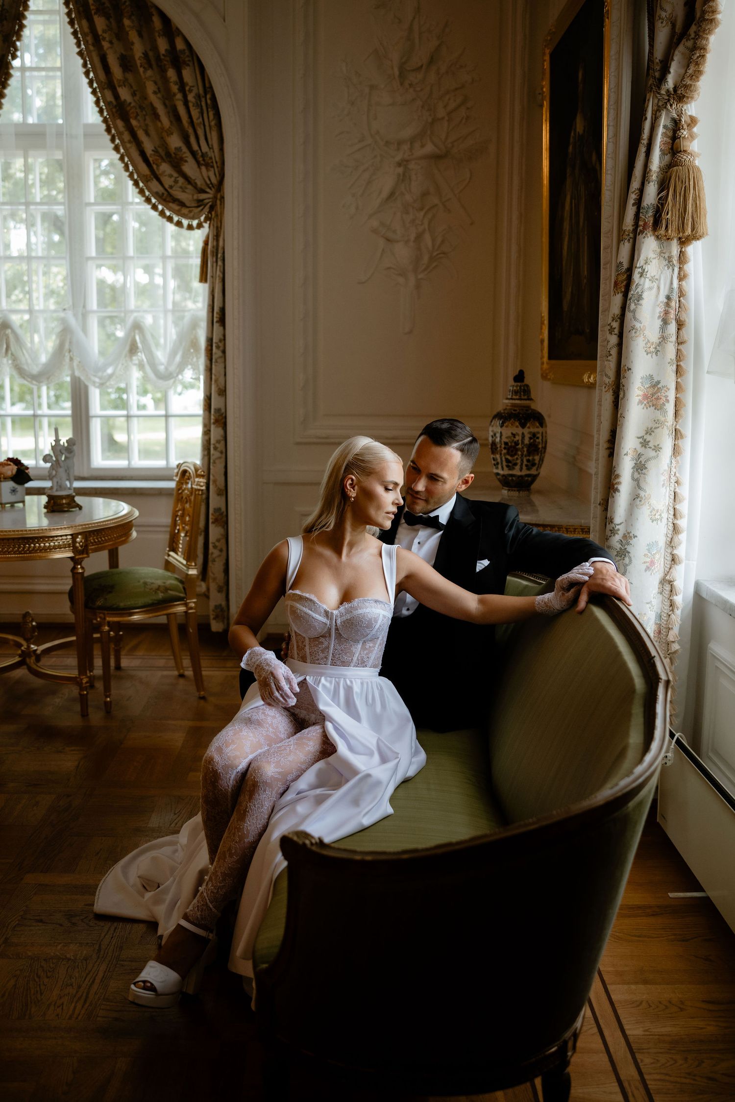 A glamorous couple in wedding attire shares a romantic moment on an antique settee by a window.