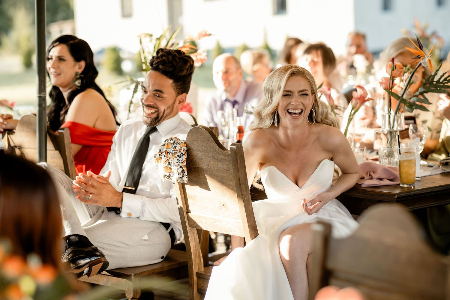 Wedding party and guests sharing laughter during an outdoor reception dinner.