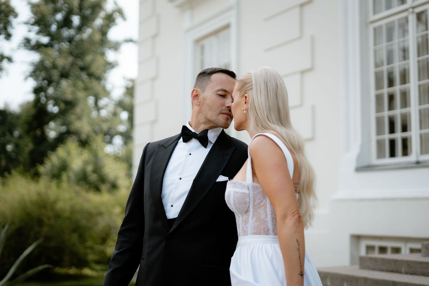 Wedding couple shares a tender moment outside a white mansion surrounded by greenery.