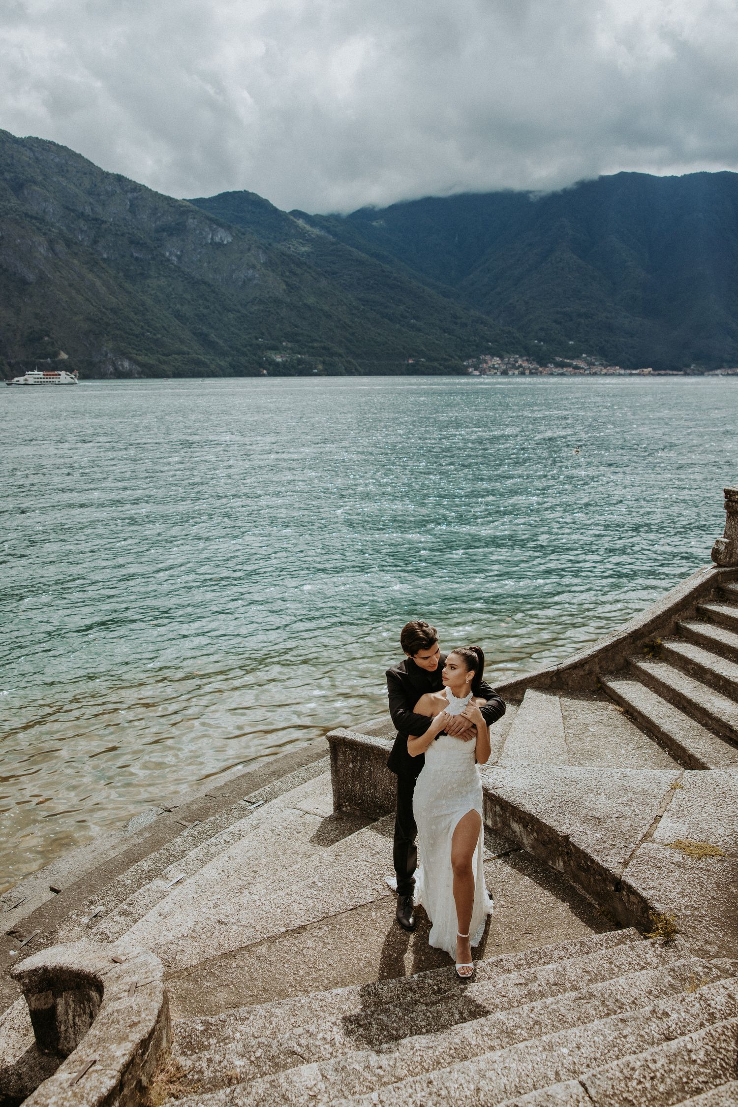 Dramatic wedding portrait on stone steps by the lake with mountain backdrop and moody sky.