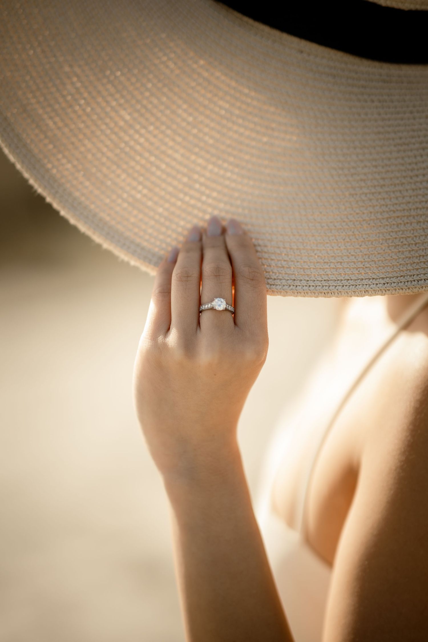 A delicate engagement ring sparkles as a hand reaches up to touch the brim of a sunhat.