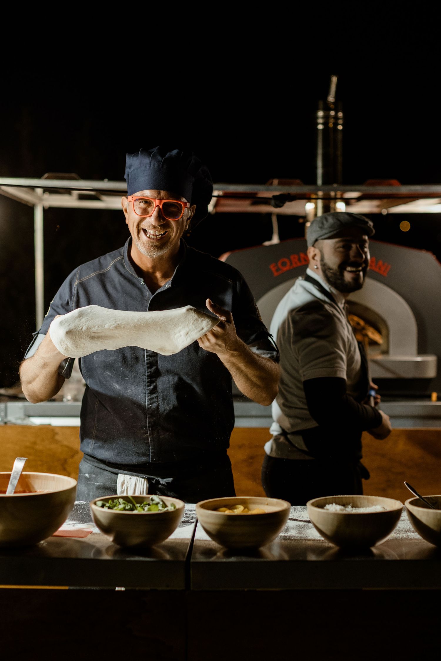 Chef in dark uniform prepares dishes in dimly lit restaurant kitchen with multiple bowls and ingredients on counter.