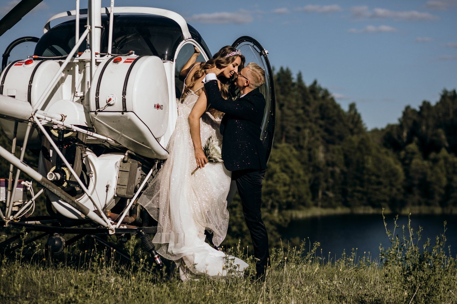 Romantic couple embraces near a small aircraft on a sunny day with trees and lake in background.