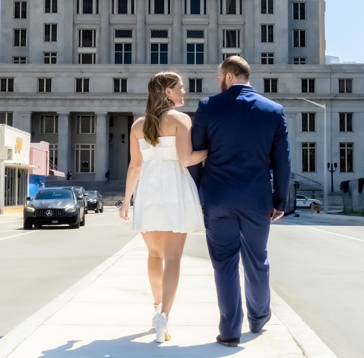 A couple in white dress and navy suit walking away from a historic building.
