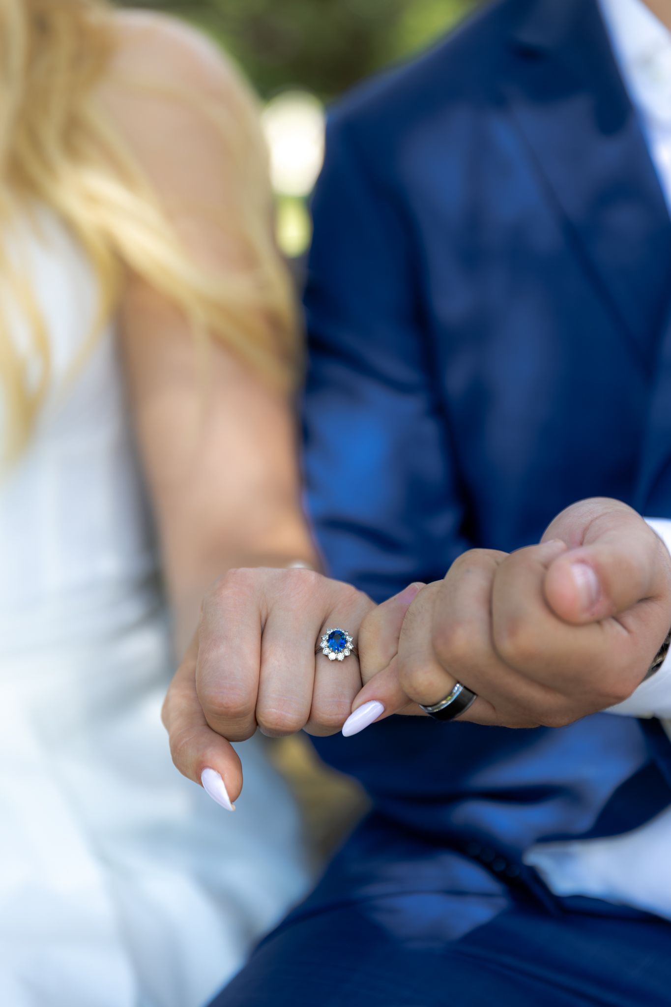 Close up shot of hands showing engagement rings with white nail polish and navy suit in soft focus background.