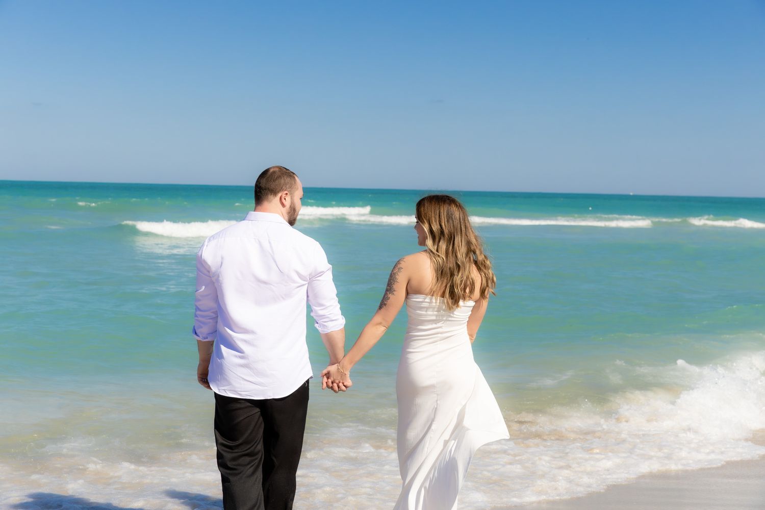 A couple holds hands while walking along a turquoise beach shore with waves crashing in the background.