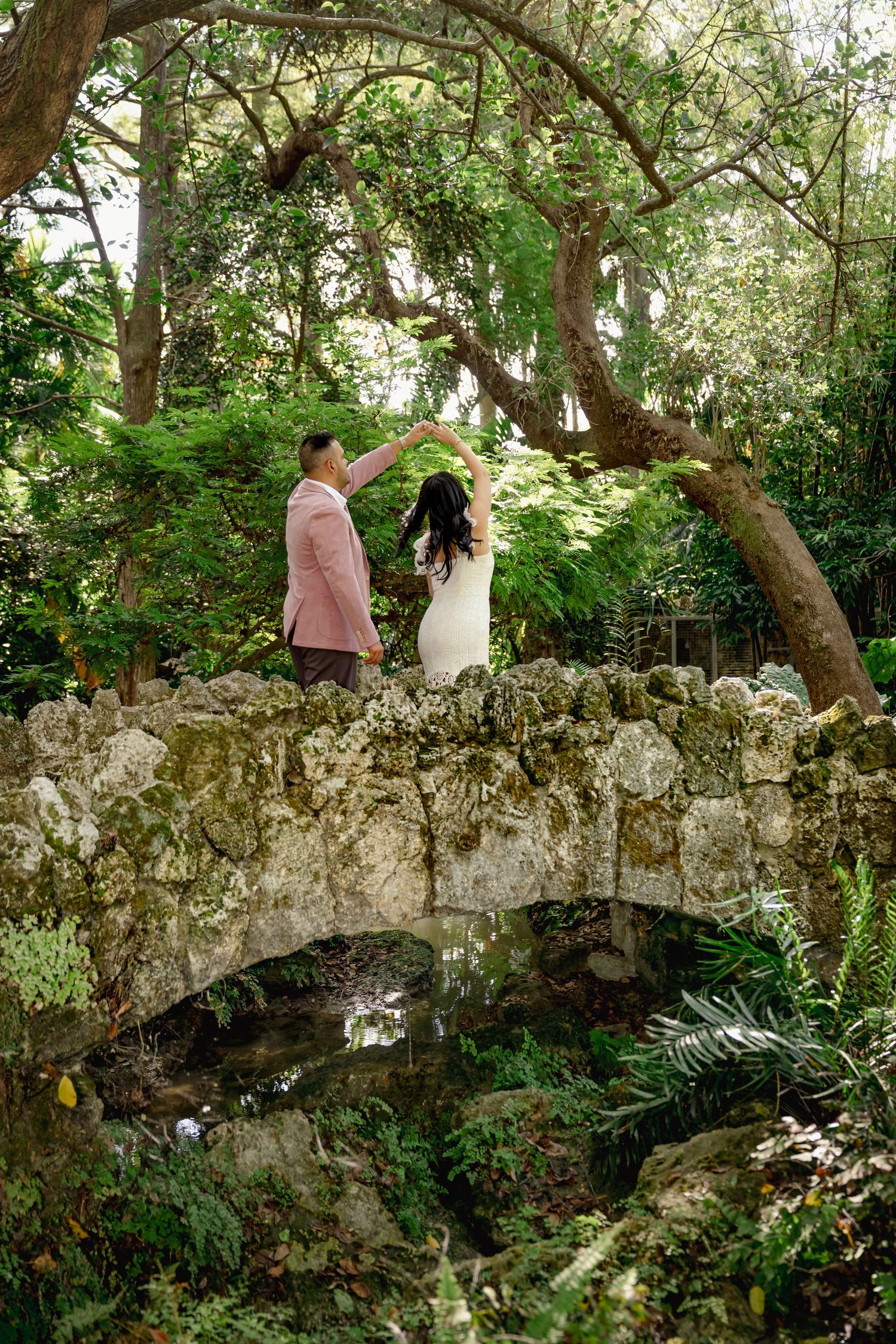 A stone bridge arches gracefully through lush green gardens with people walking across the decorative pathway.