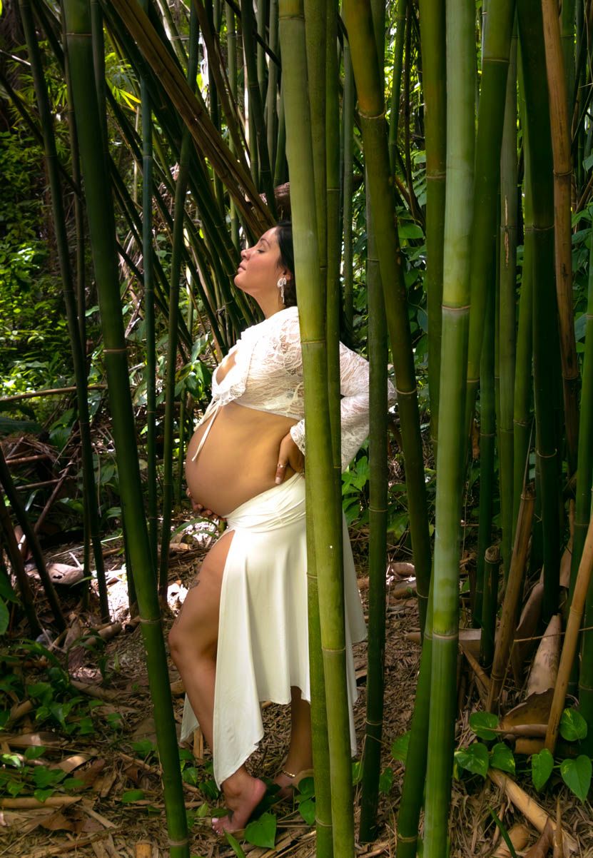 An artistic maternity photo among tall green bamboo stalks showcases a flowing white dress.