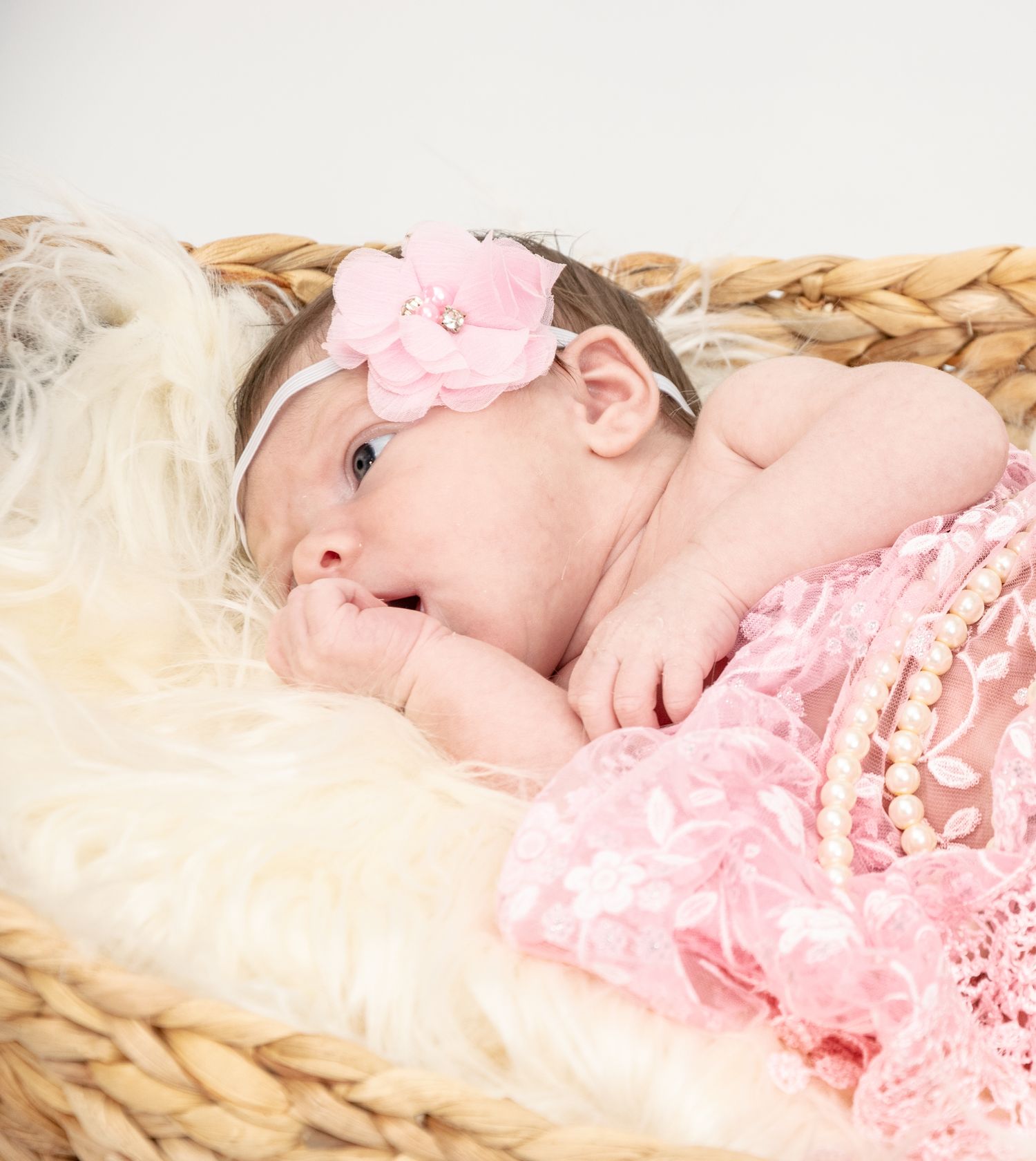 A peaceful newborn rests in a woven basket adorned with pink accessories and lace.