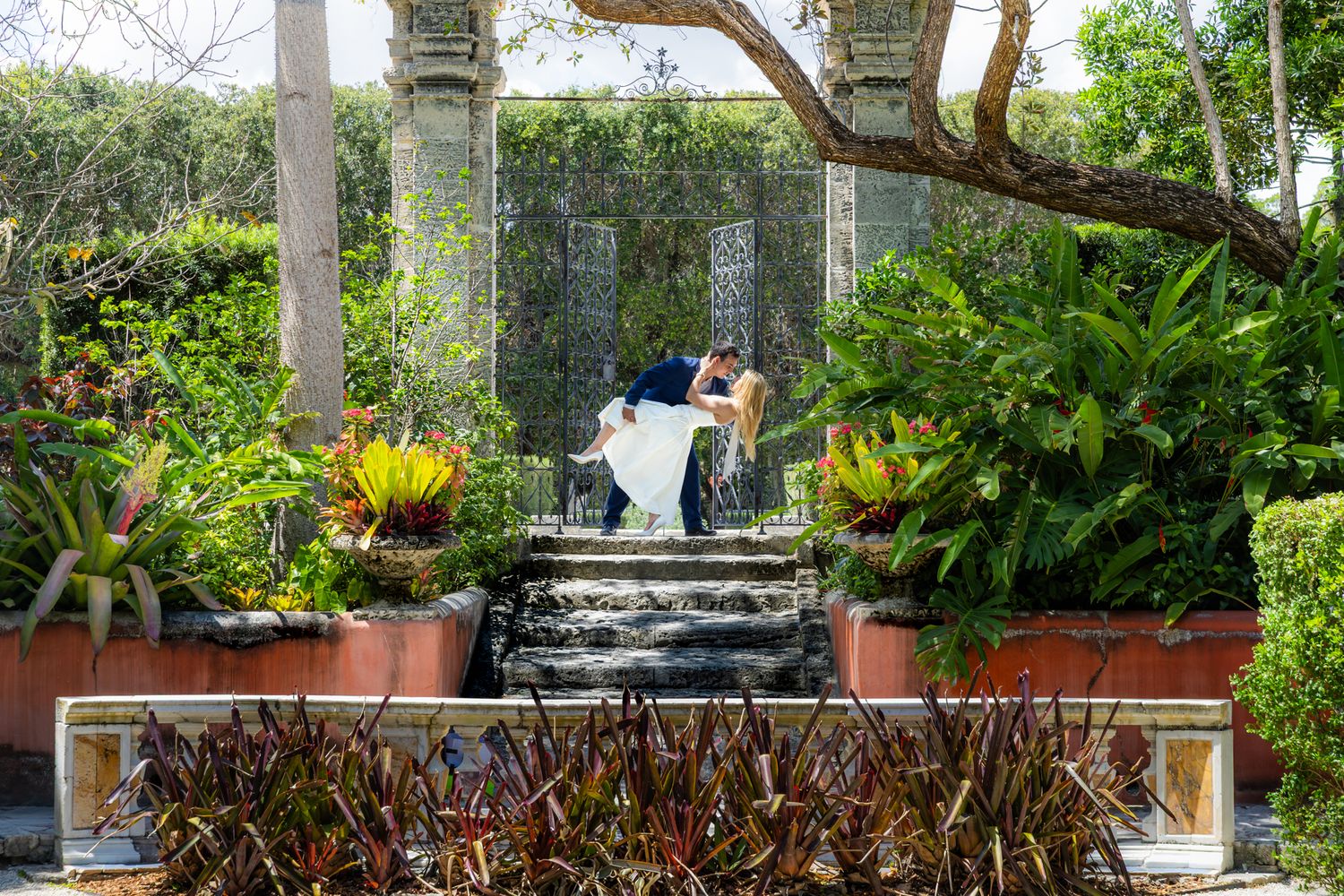 Ornate garden terrace with potted plants and stone steps leading to a seating area surrounded by lush tropical foliage.