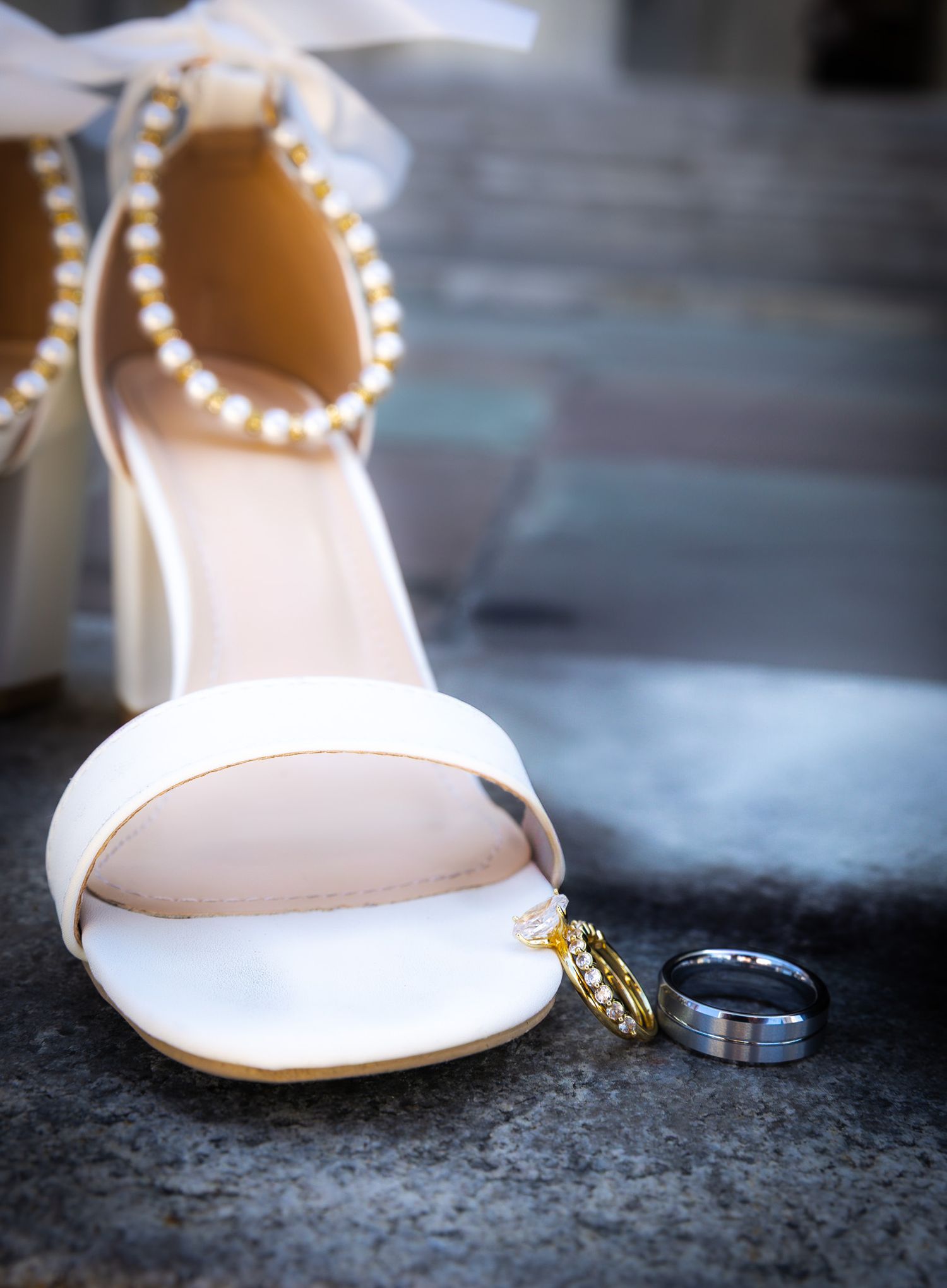 White wedding shoe with pearl ankle strap detail alongside wedding rings on dark surface.