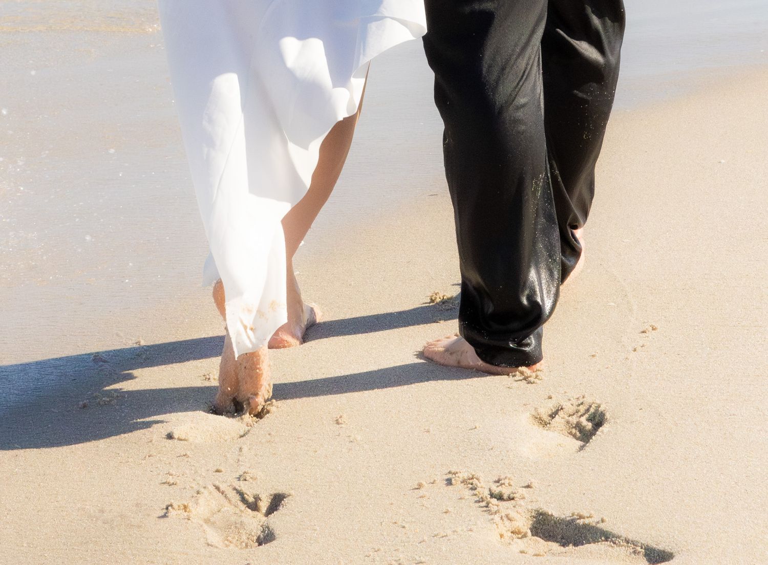Two people walking barefoot on a sandy beach leaving footprints in their wake during bright daylight.