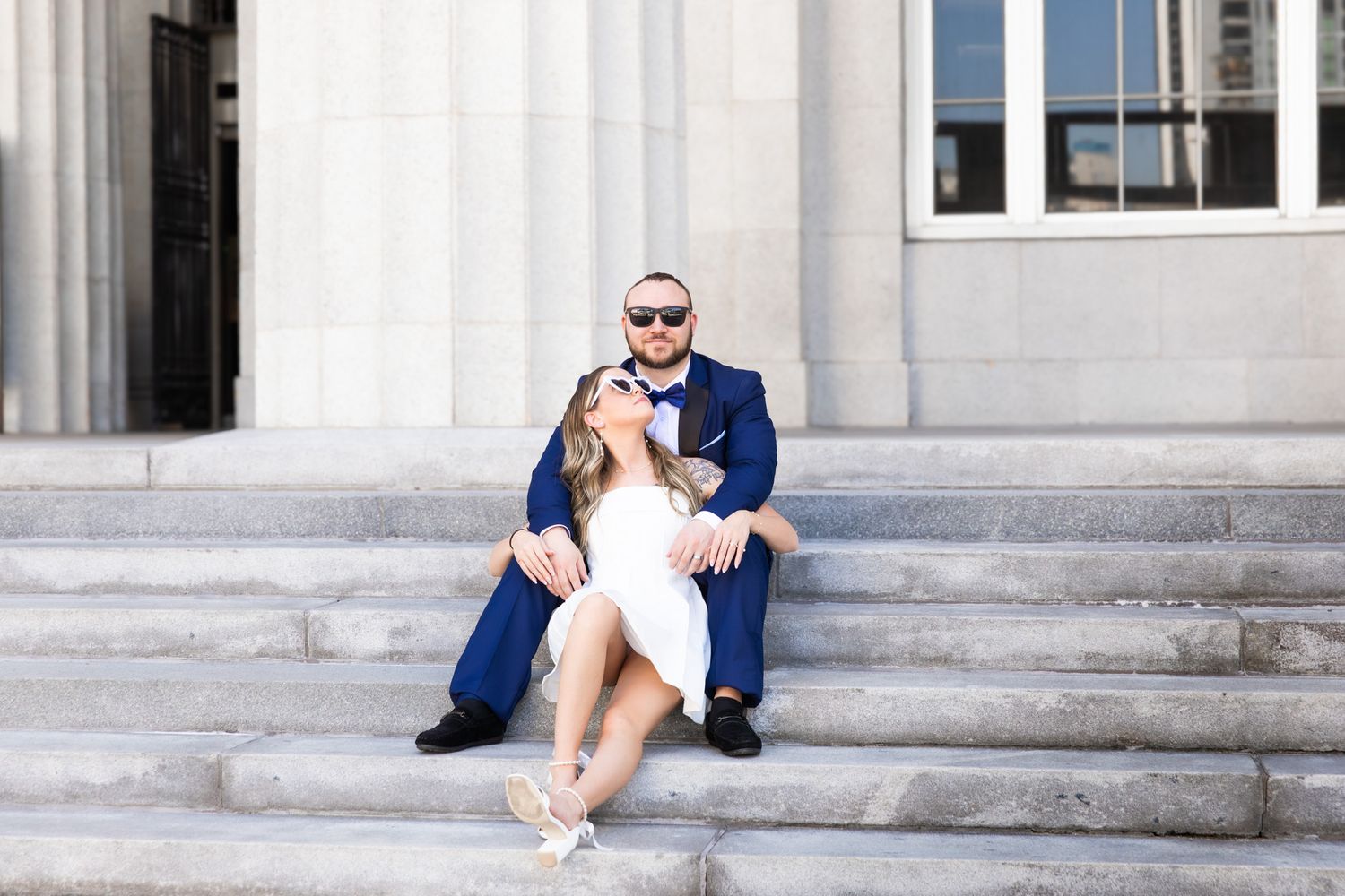 A couple in navy and white attire sitting on stone steps of a historic building with columns.