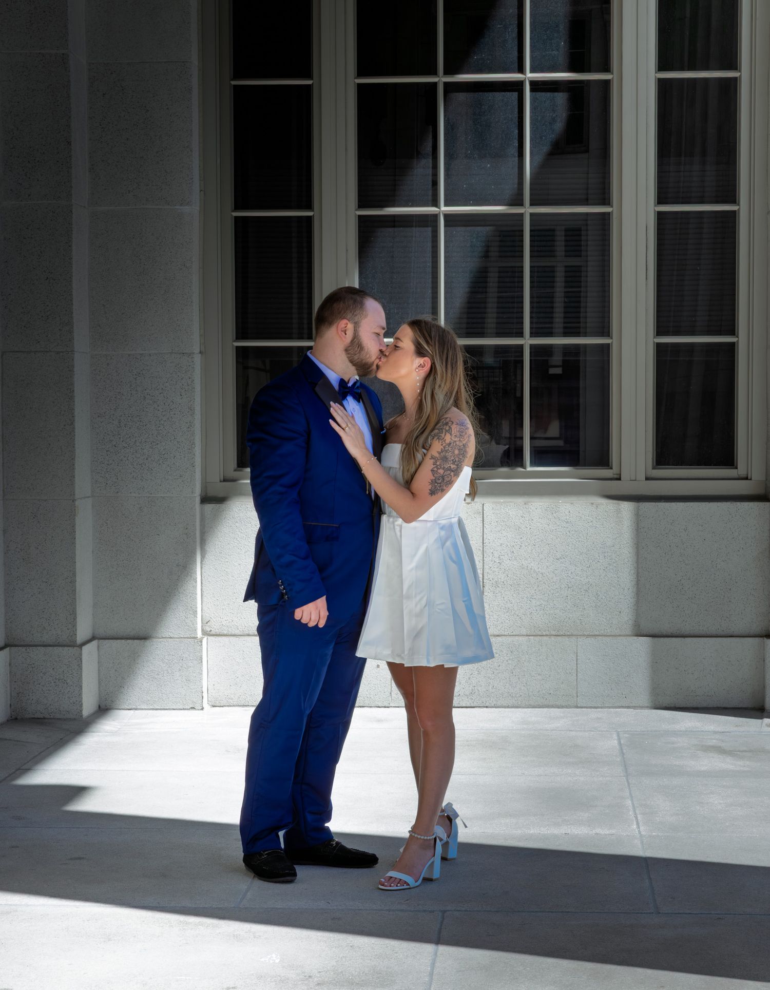 Romantic couple in formal attire share intimate moment against sunlit classical building.