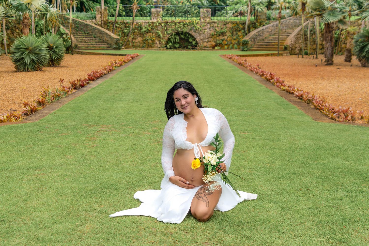 A maternity photo on a manicured lawn with flower borders shows a flowing white dress and sunlit pose.