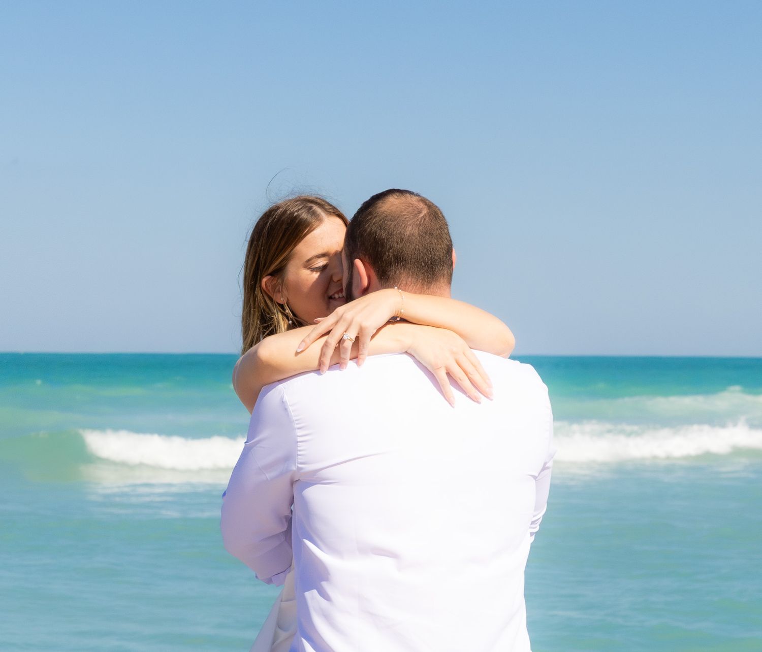 Two people embrace in a romantic moment on a beach with turquoise waters and clear blue sky in the background.