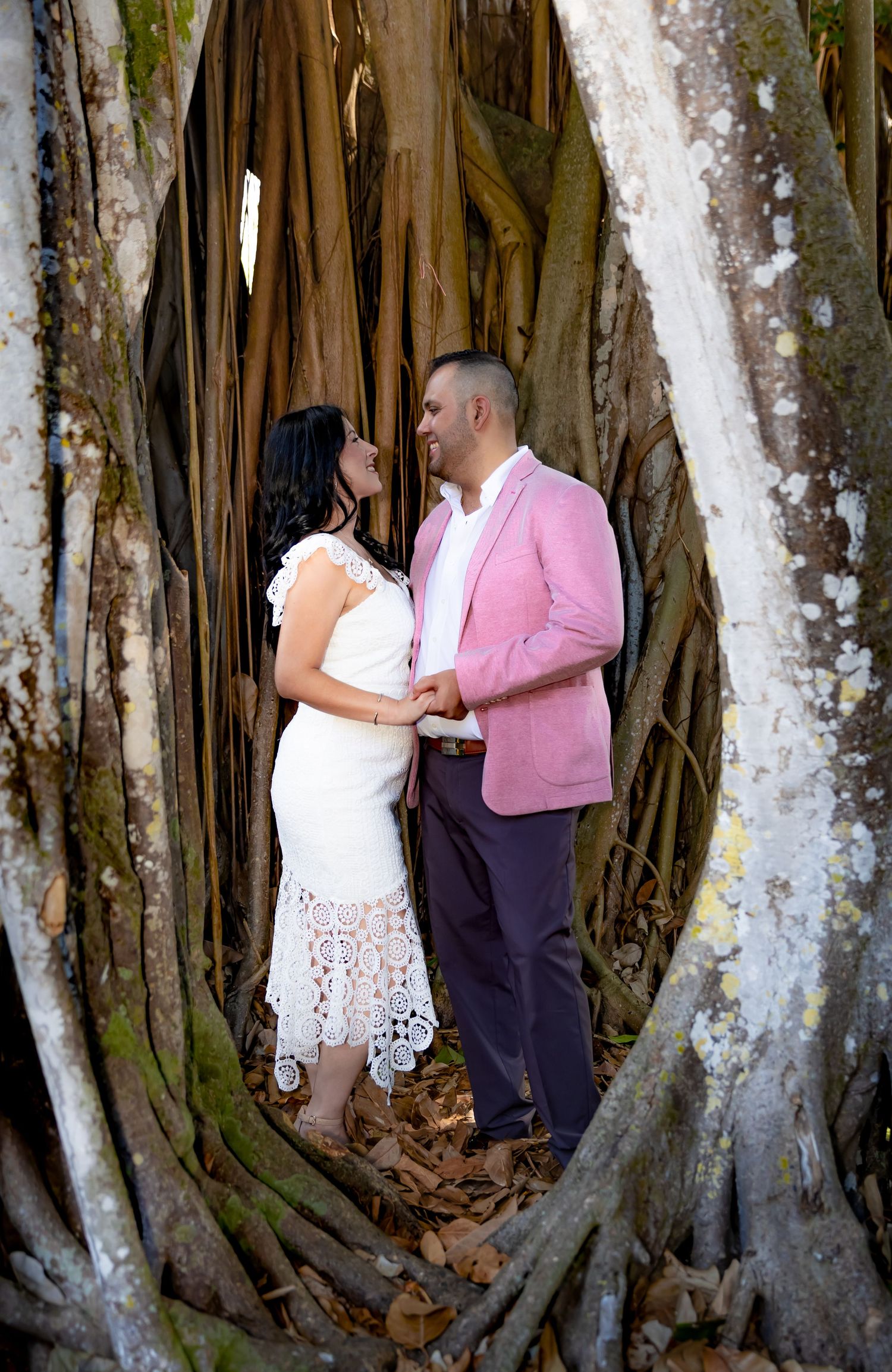 A romantic couple poses between large banyan tree roots, with the person in a pink blazer and white dress creating contrast.