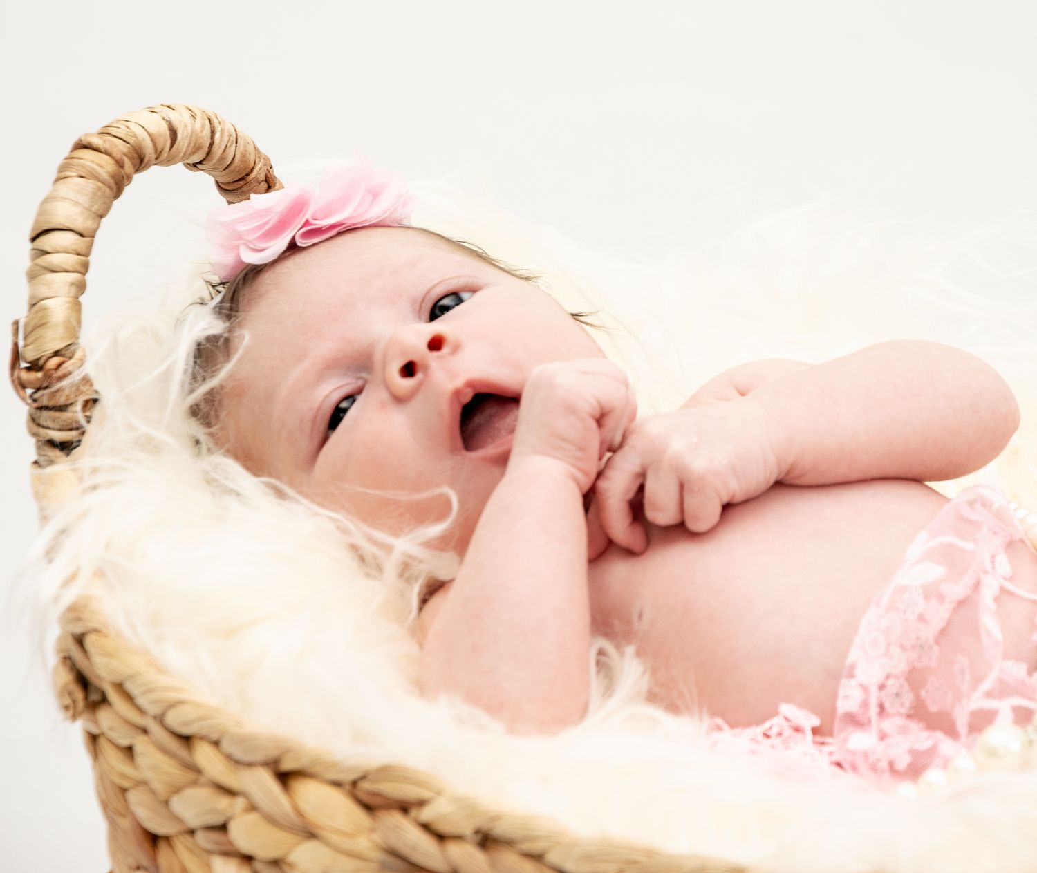 A smiling newborn wearing a pink headband lies in a woven basket with cream-colored lining.