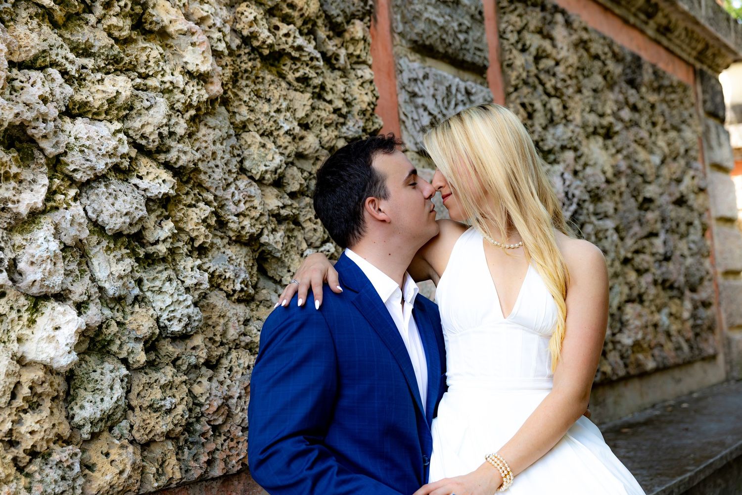 A couple in wedding attire share a romantic moment against a textured stone wall on their special day.