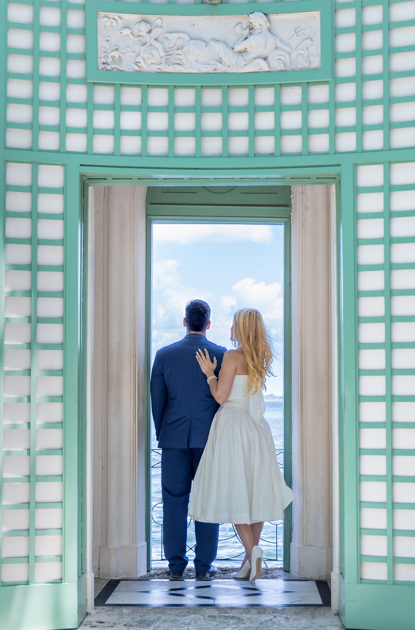 A couple stands in a mint green doorway overlooking a scenic balcony view, with classic architectural details surrounding them.