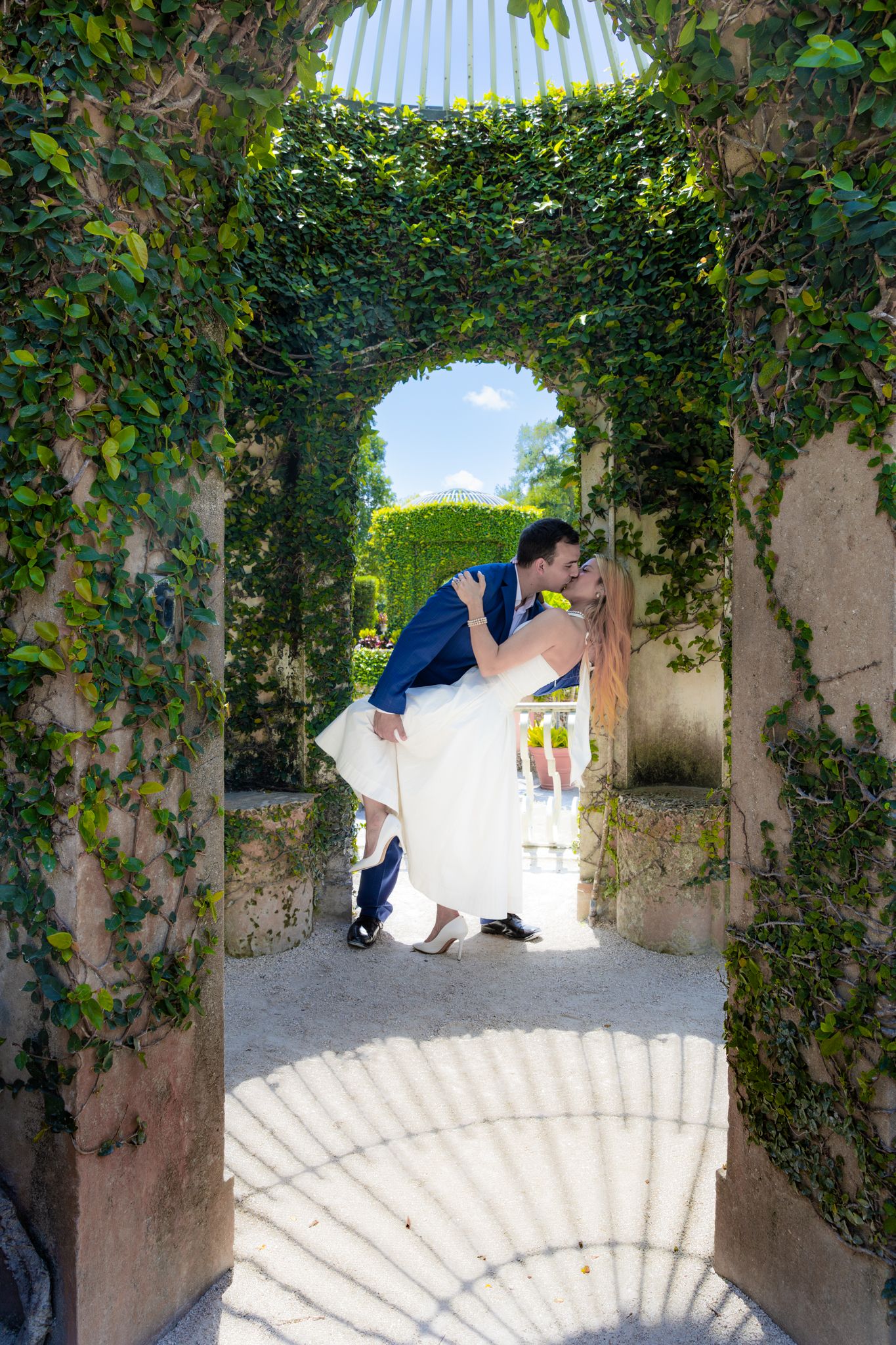 A romantic couple shares an intimate moment under a vine-covered archway in an elegant garden setting.