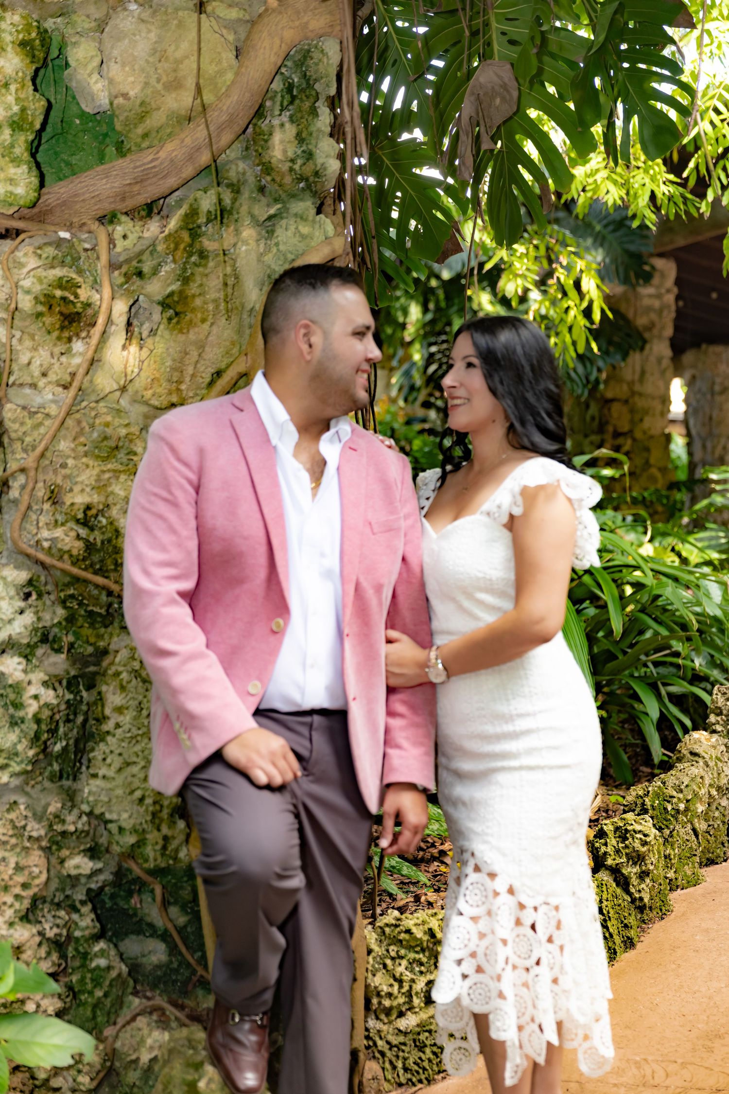 A couple in pink and white attire share romantic moments among lush tropical foliage during their outdoor photo session.
