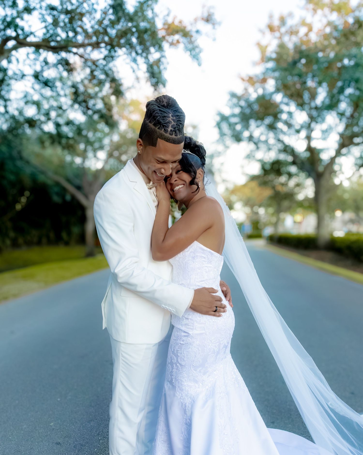 Romantic wedding couple embrace on tree-lined road in all-white attire.