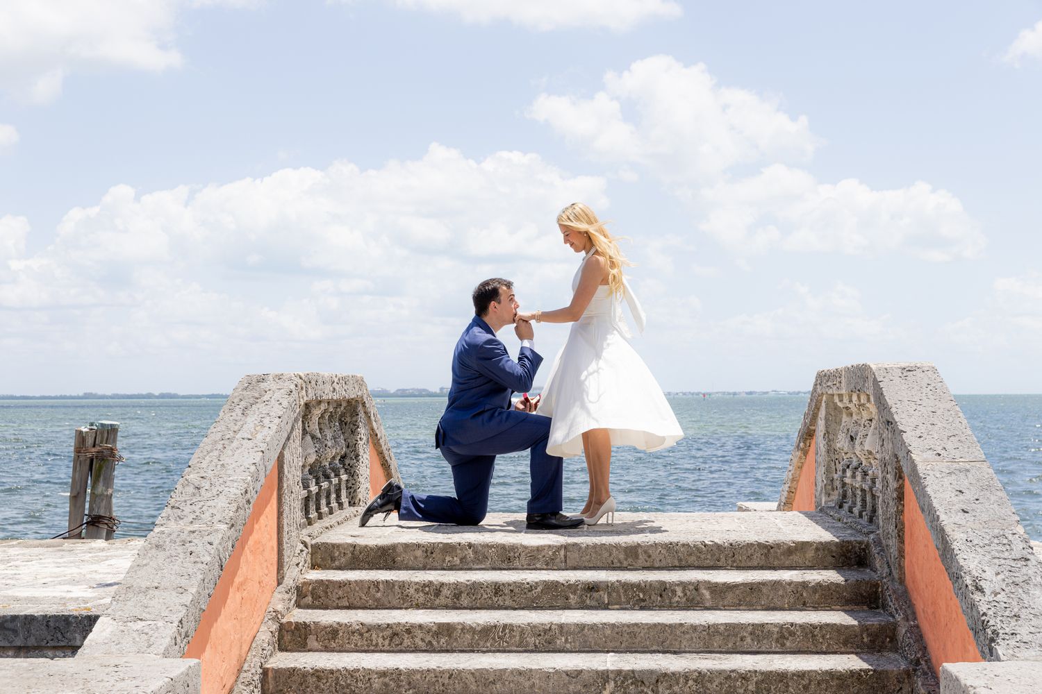 A romantic proposal scene with couple on coastal steps overlooking the ocean on a bright sunny day.