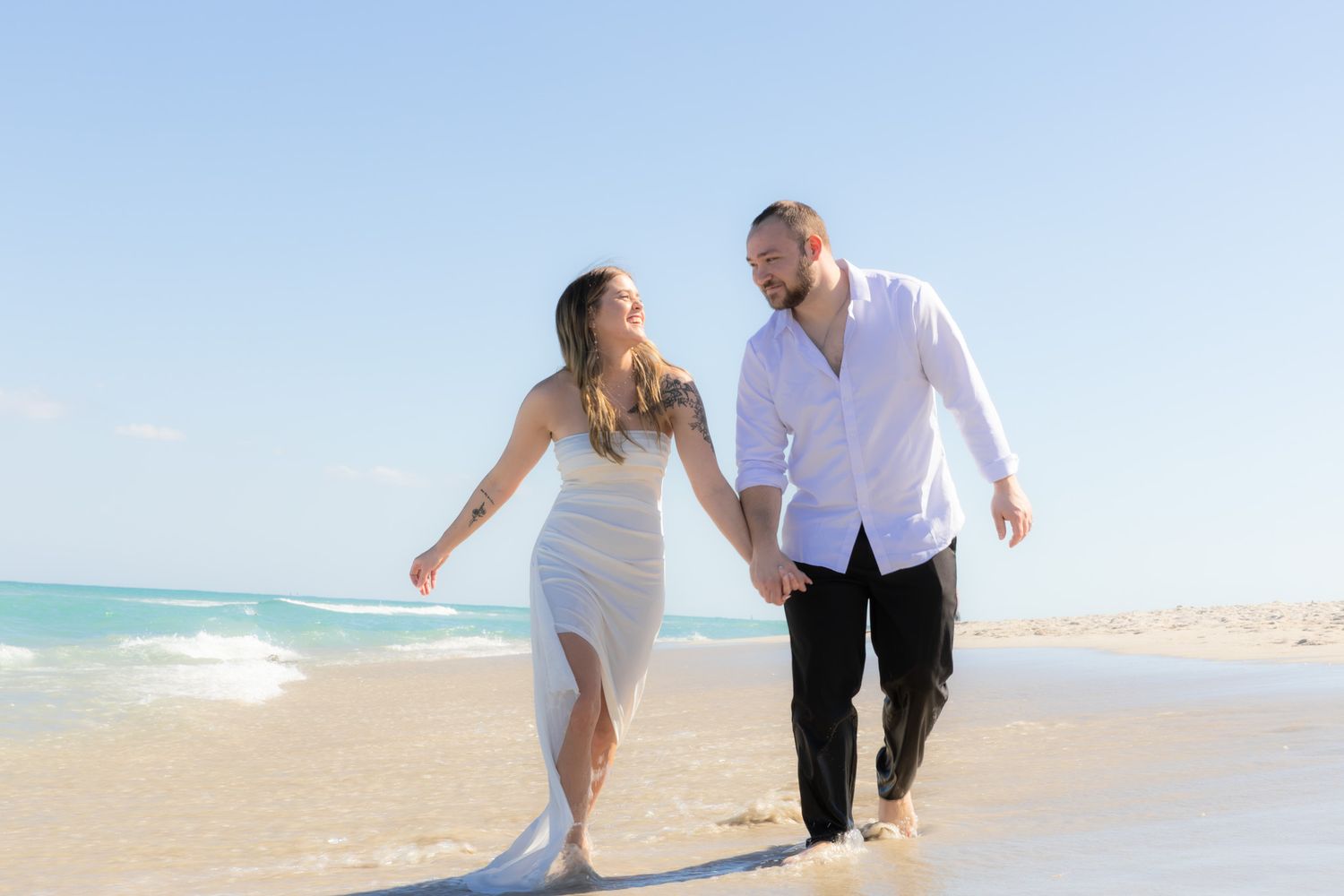 Couple in white outfits walking joyfully along a tropical beach with turquoise ocean waters and clear blue sky.