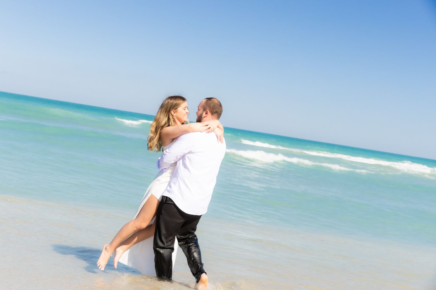 A romantic couple embraces on a pristine beach with turquoise water and clear blue skies in the background.
