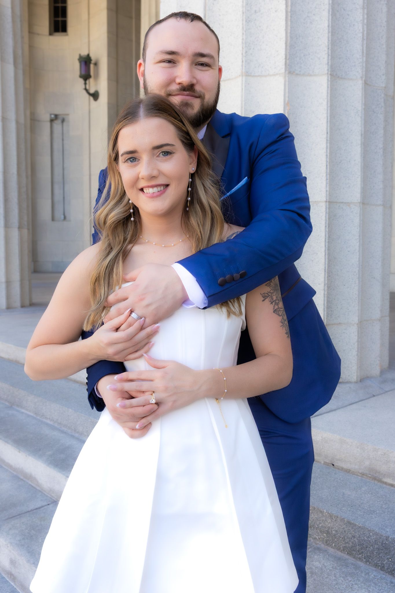 Formal portrait of couple in blue suit and white dress against classical architecture.