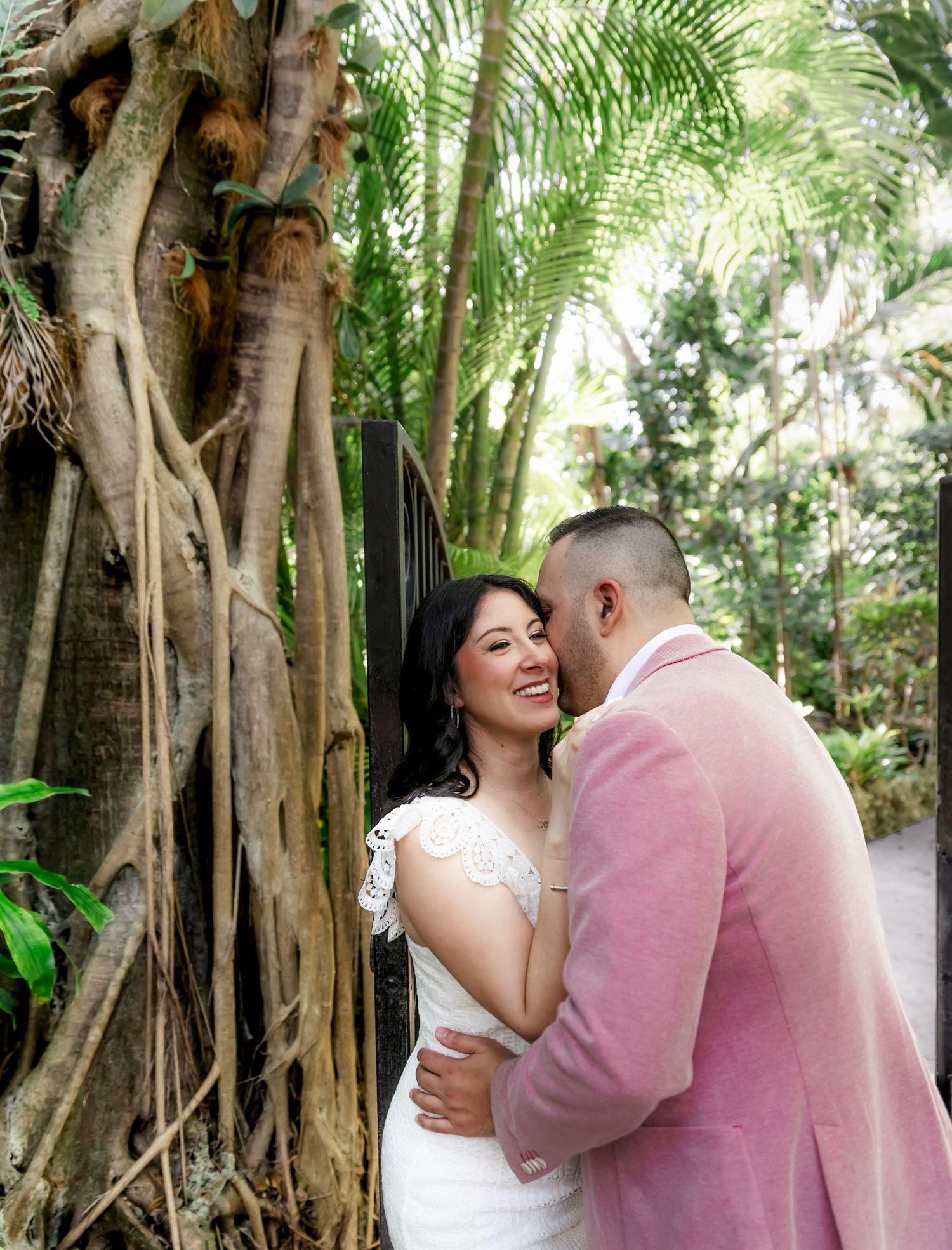 Romantic couple in wedding attire share tender embraces in a tropical garden setting with lush palm trees.