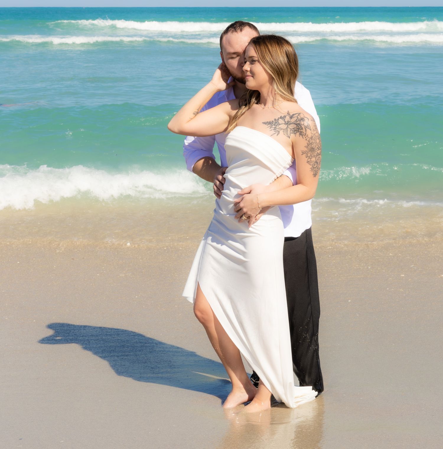 A couple embraces and kisses on a sunny beach with turquoise ocean waves in the background.