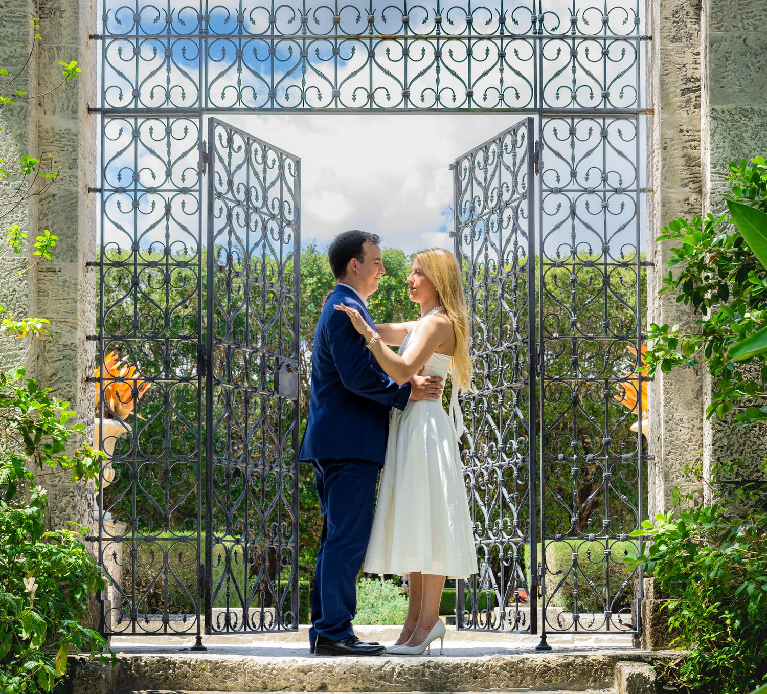 A couple shares a romantic moment in front of an ornate wrought iron gate surrounded by blooming garden flowers.