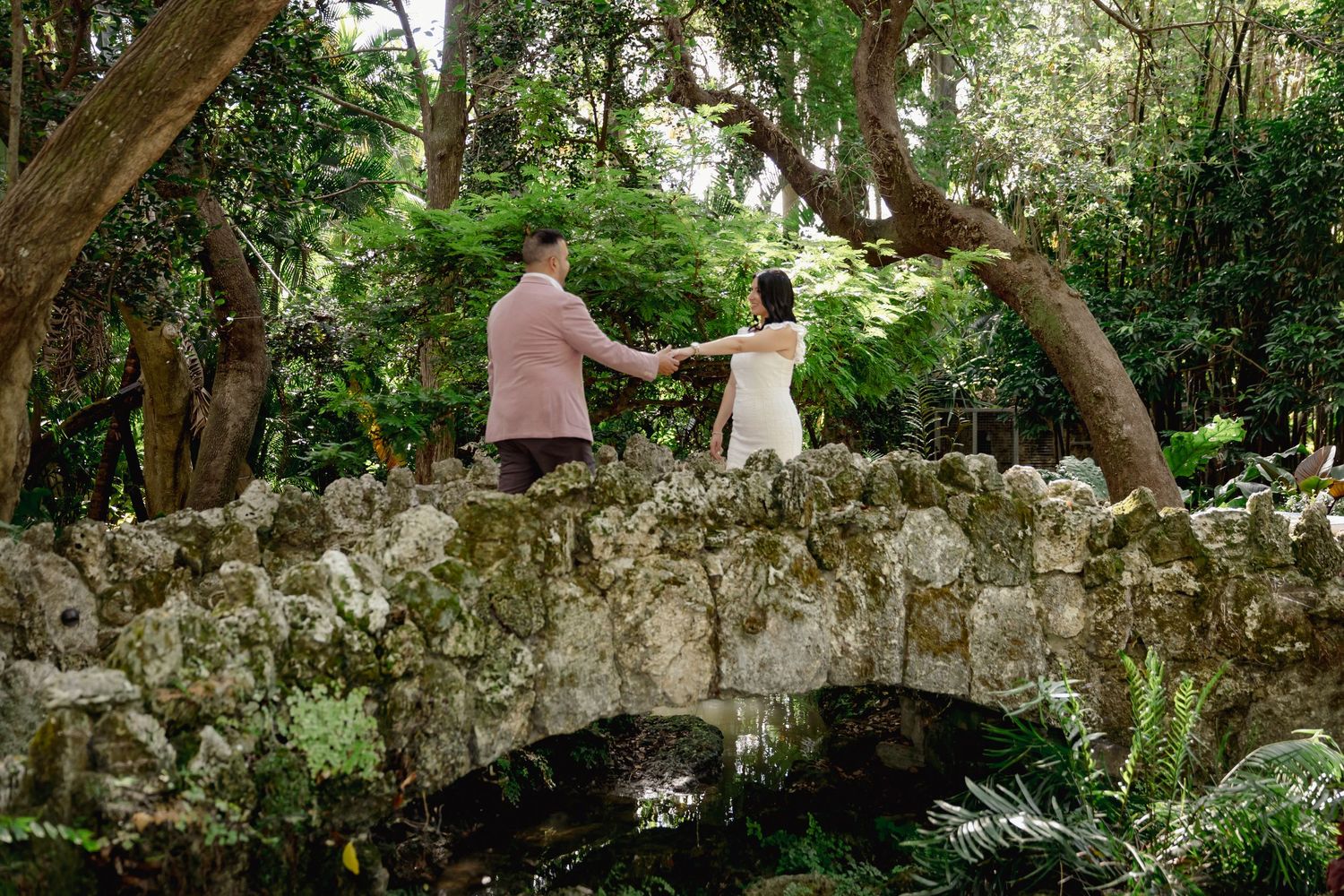 Two people sit on an aged stone wall surrounded by lush green forest canopy in a peaceful outdoor setting.