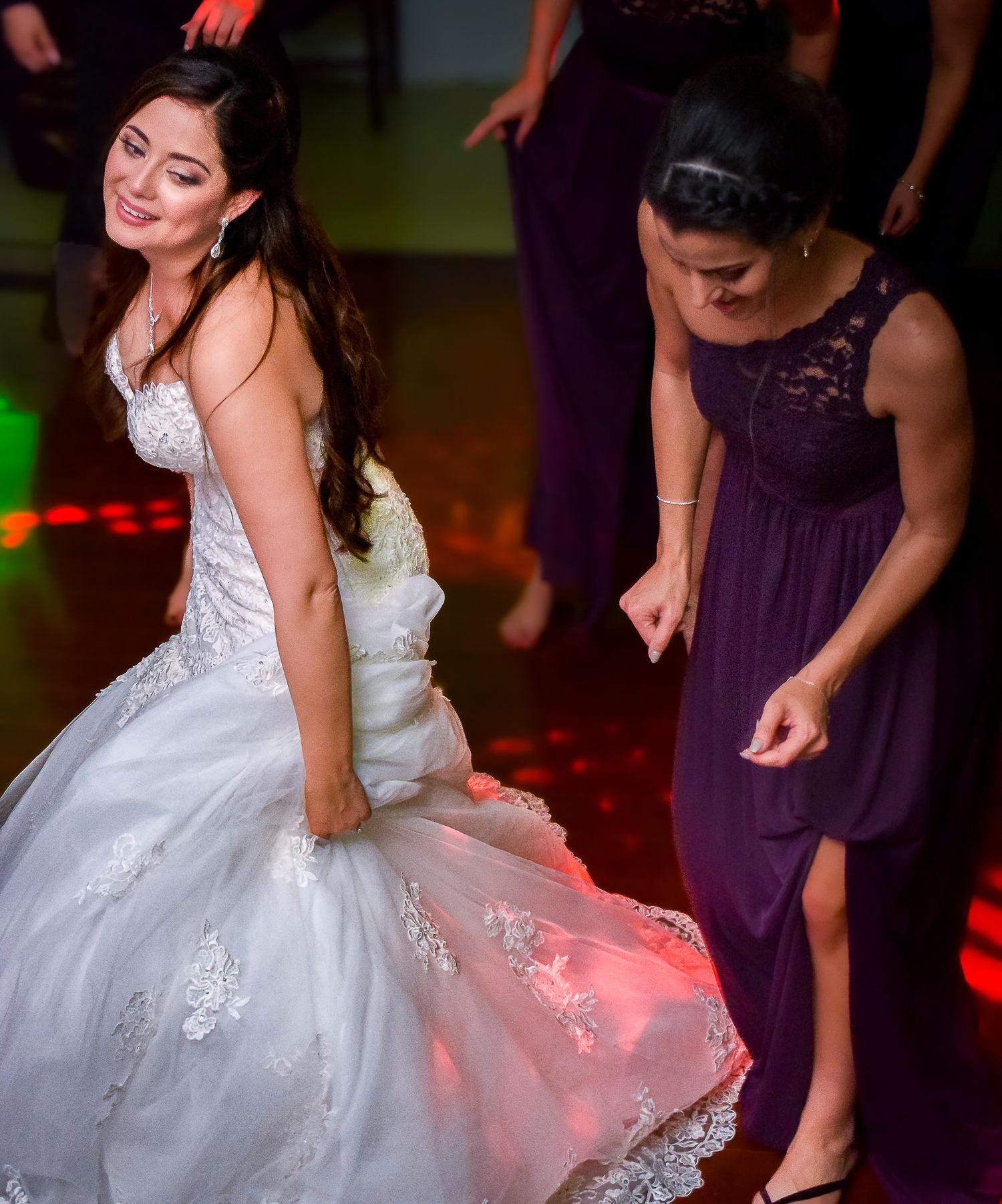 Bride in white lace gown dancing on illuminated dance floor with bridesmaid in purple dress.