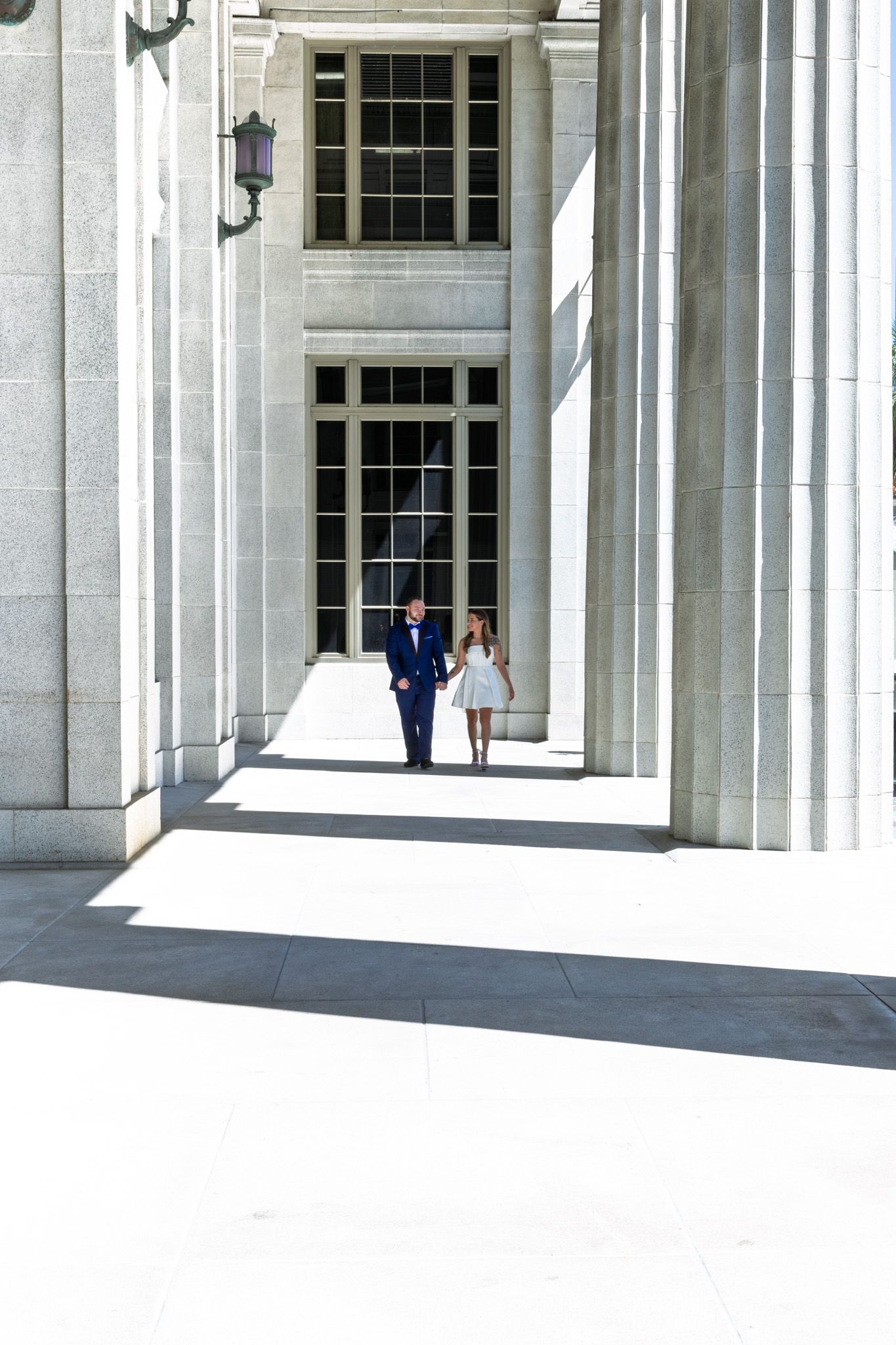 A couple walking between white columns of a grand neoclassical building entrance with tall windows.