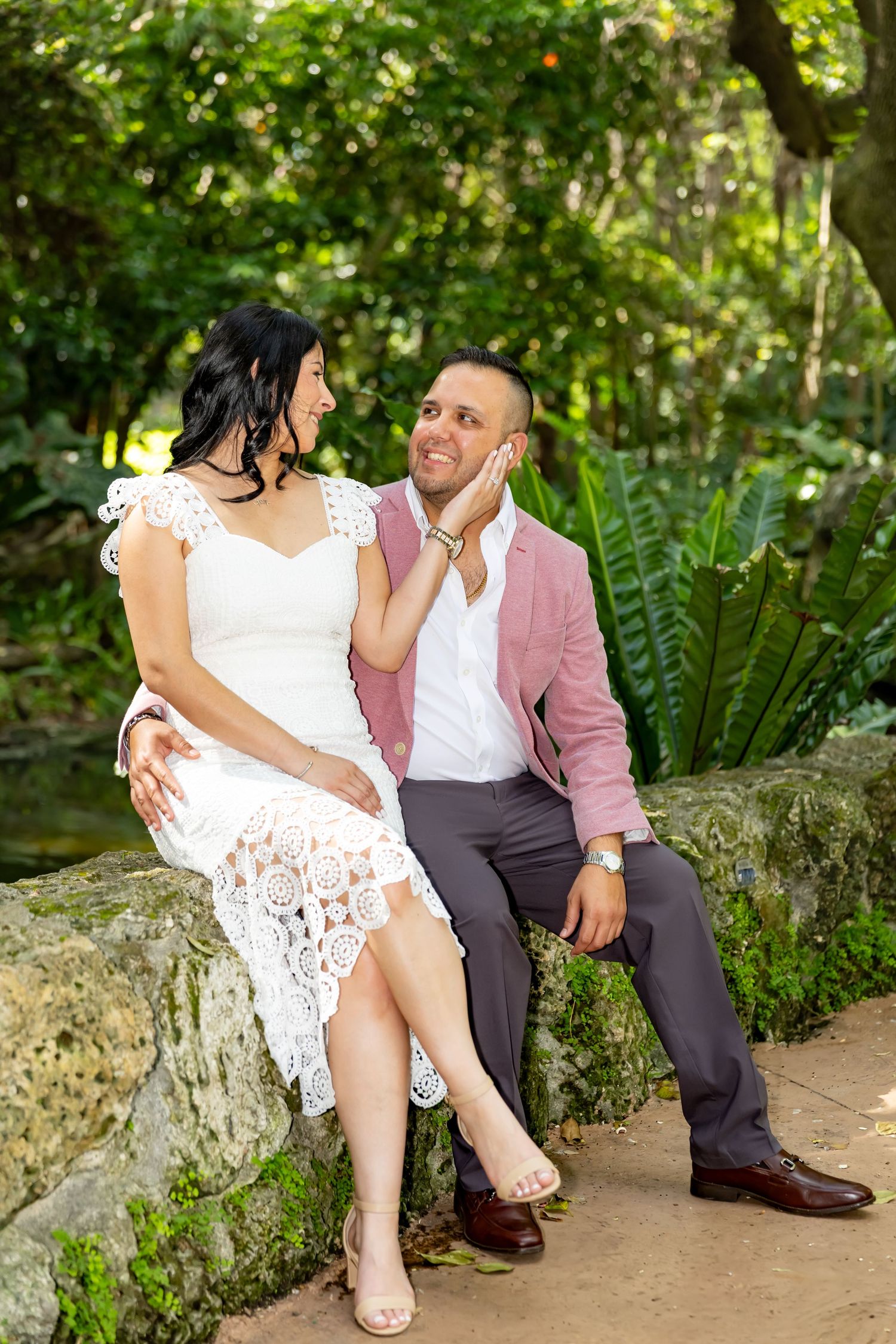 Couple sits on stone wall in garden setting with lush greenery, bride wearing white lace dress and groom in pink blazer.