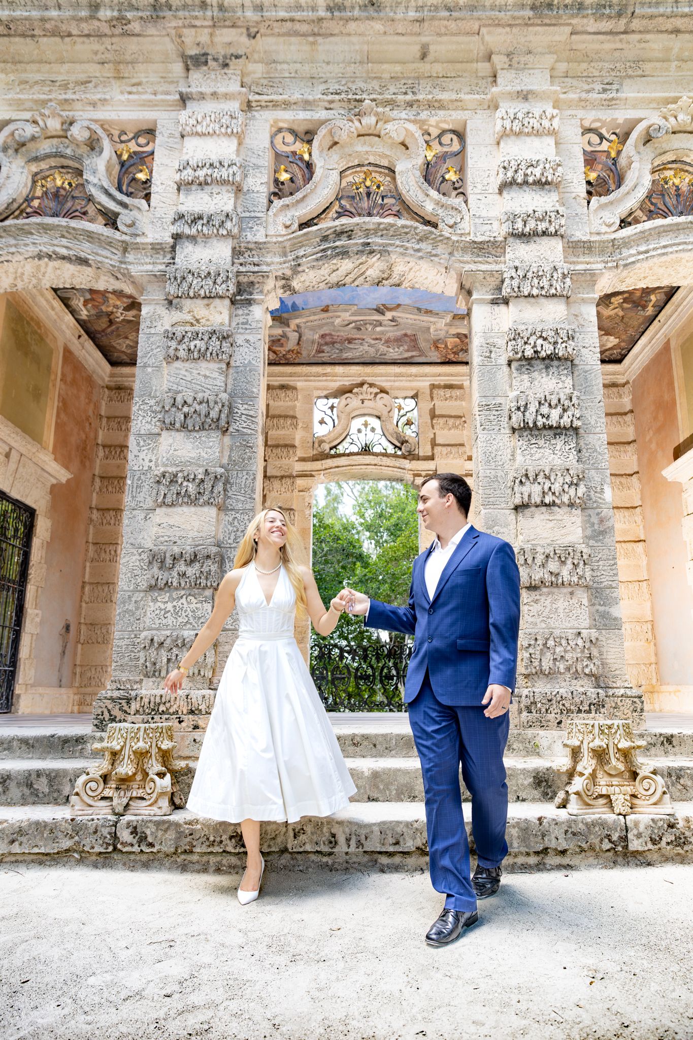 Newlywed couple poses under ornate stone archway with carved pillars and classical architecture at elegant garden venue.