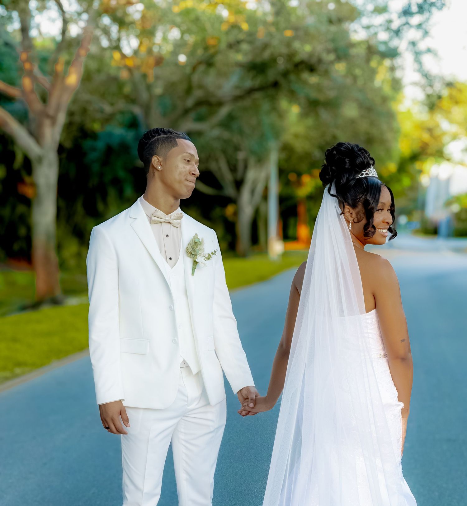 A newlywed couple walks hand in hand down a tree-lined street.