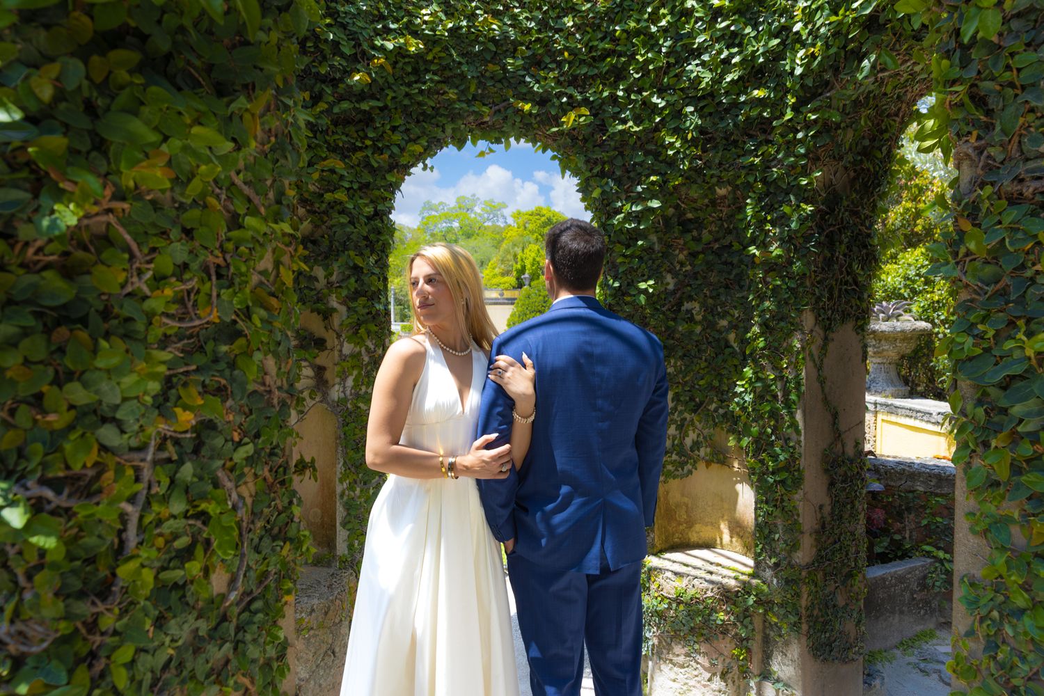 A couple in wedding attire stands beneath a lush green archway in a garden setting on a sunny day.