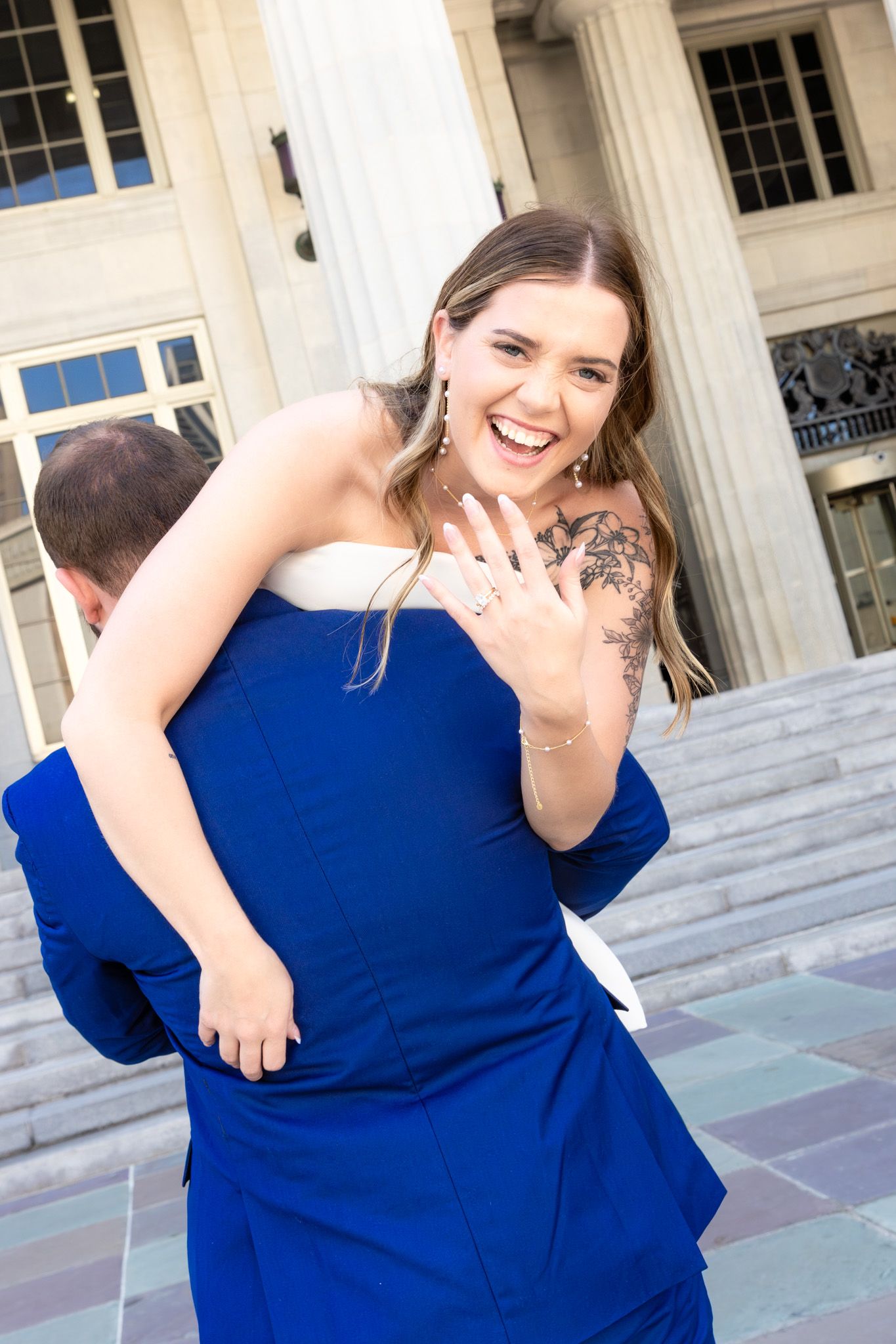 A person in blue showing off an engagement ring in front of a columned building.