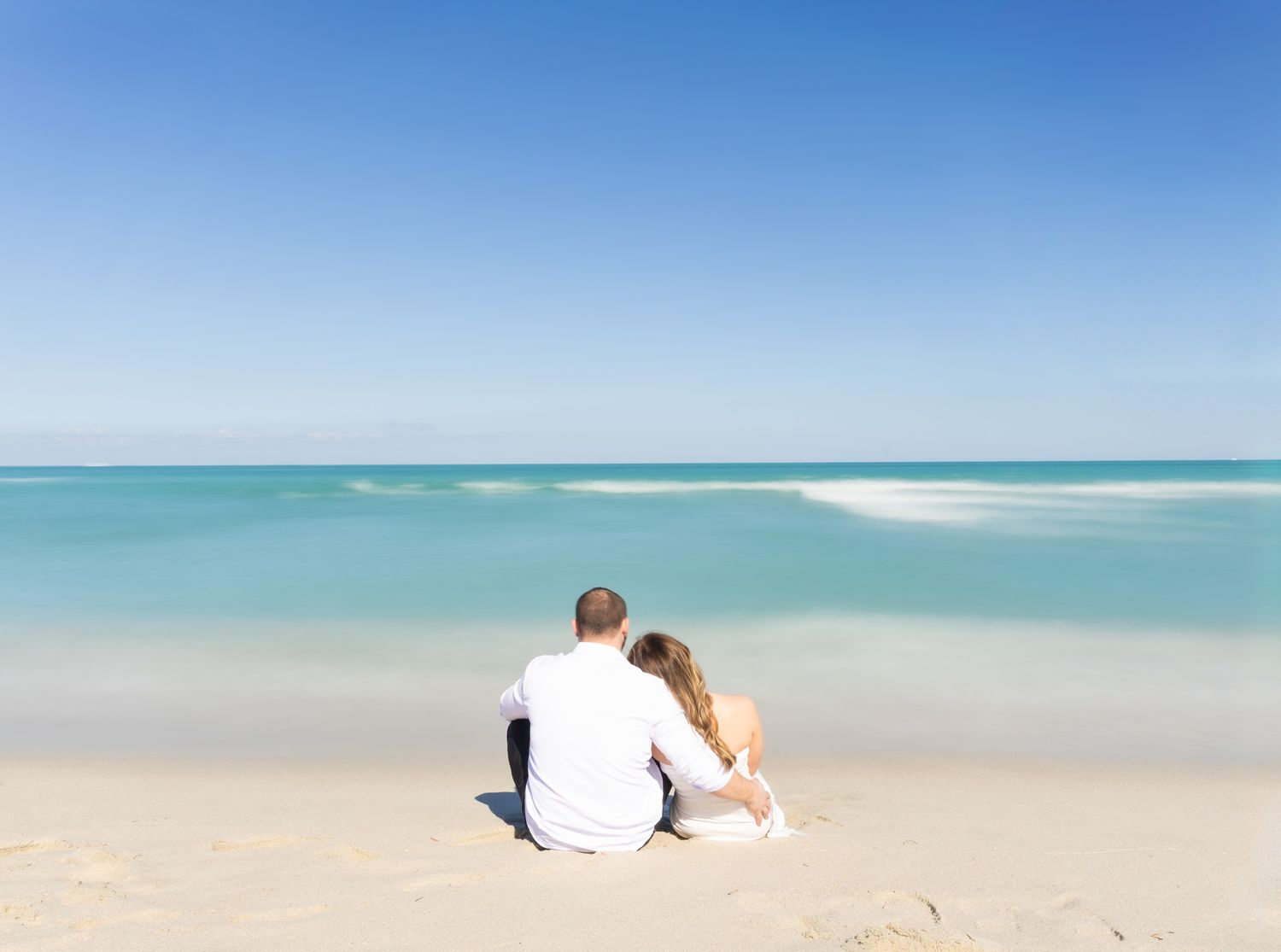 A couple sits together on a pristine white sand beach overlooking turquoise ocean waters on a sunny day.