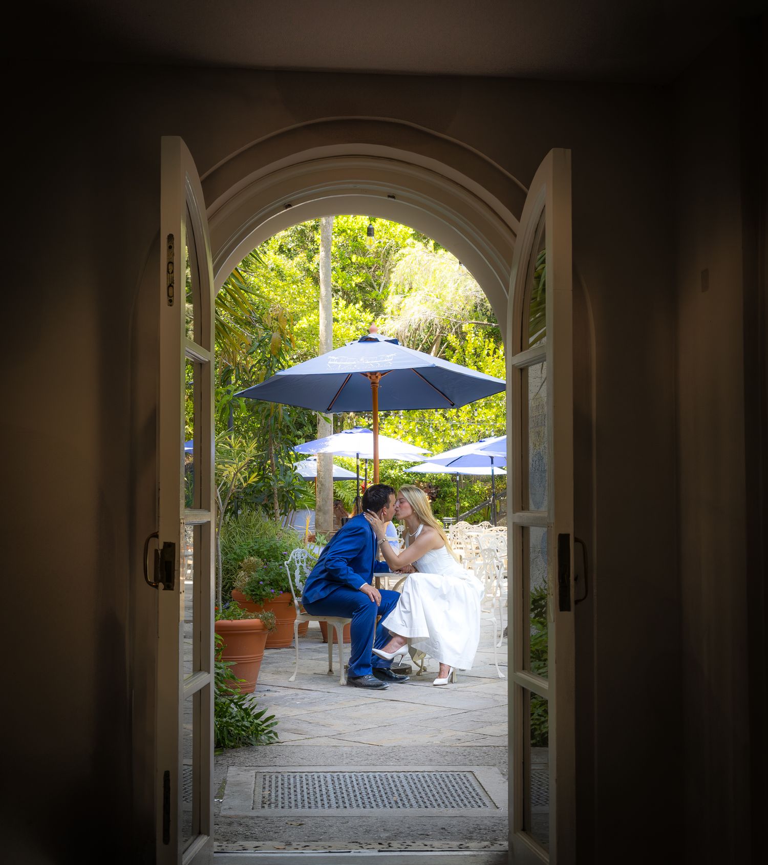 View through an arched doorway of two people seated at a table with blue umbrellas in a sunny garden patio.