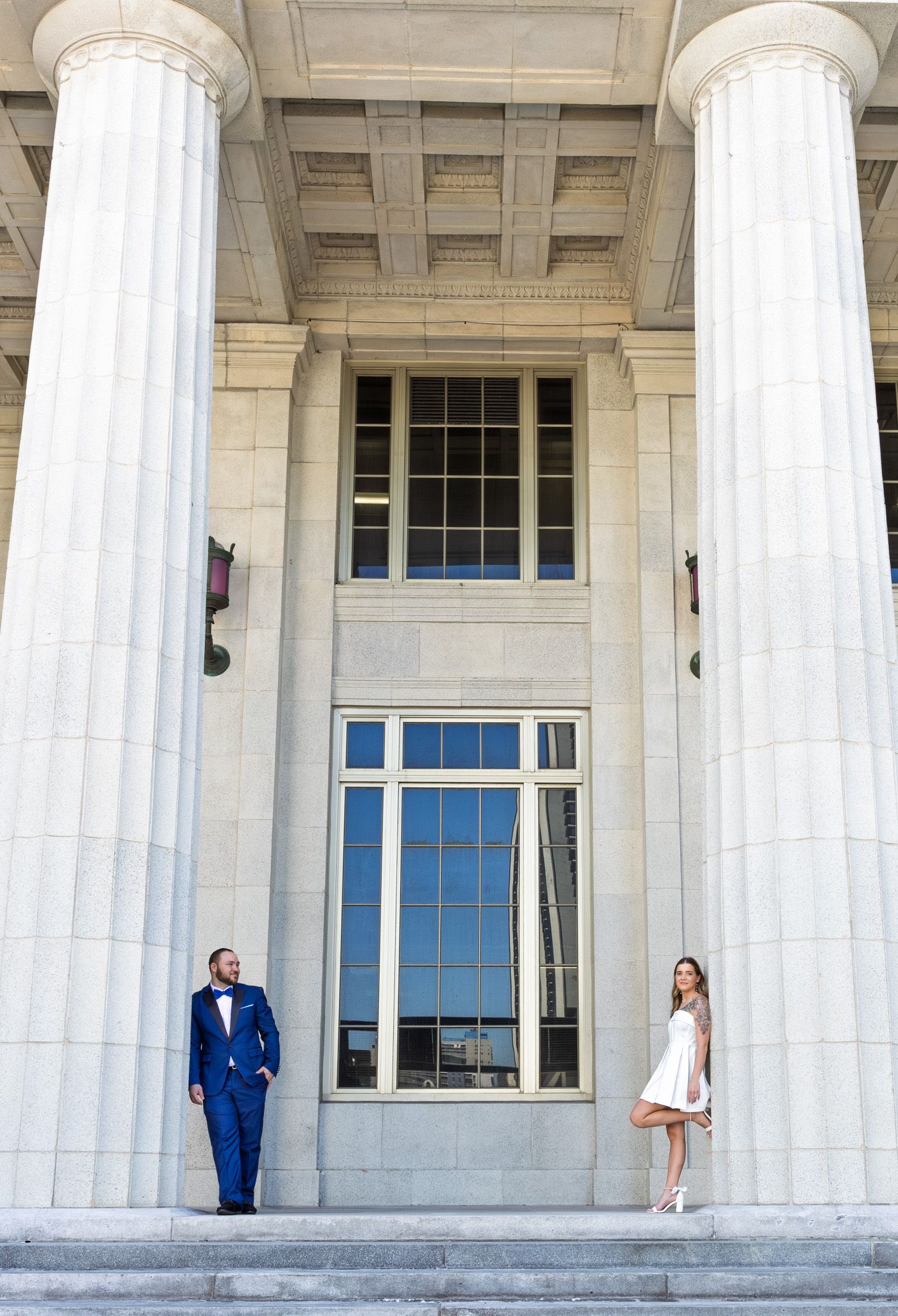 A couple posing between tall white columns at opposite ends of a grand building entrance.